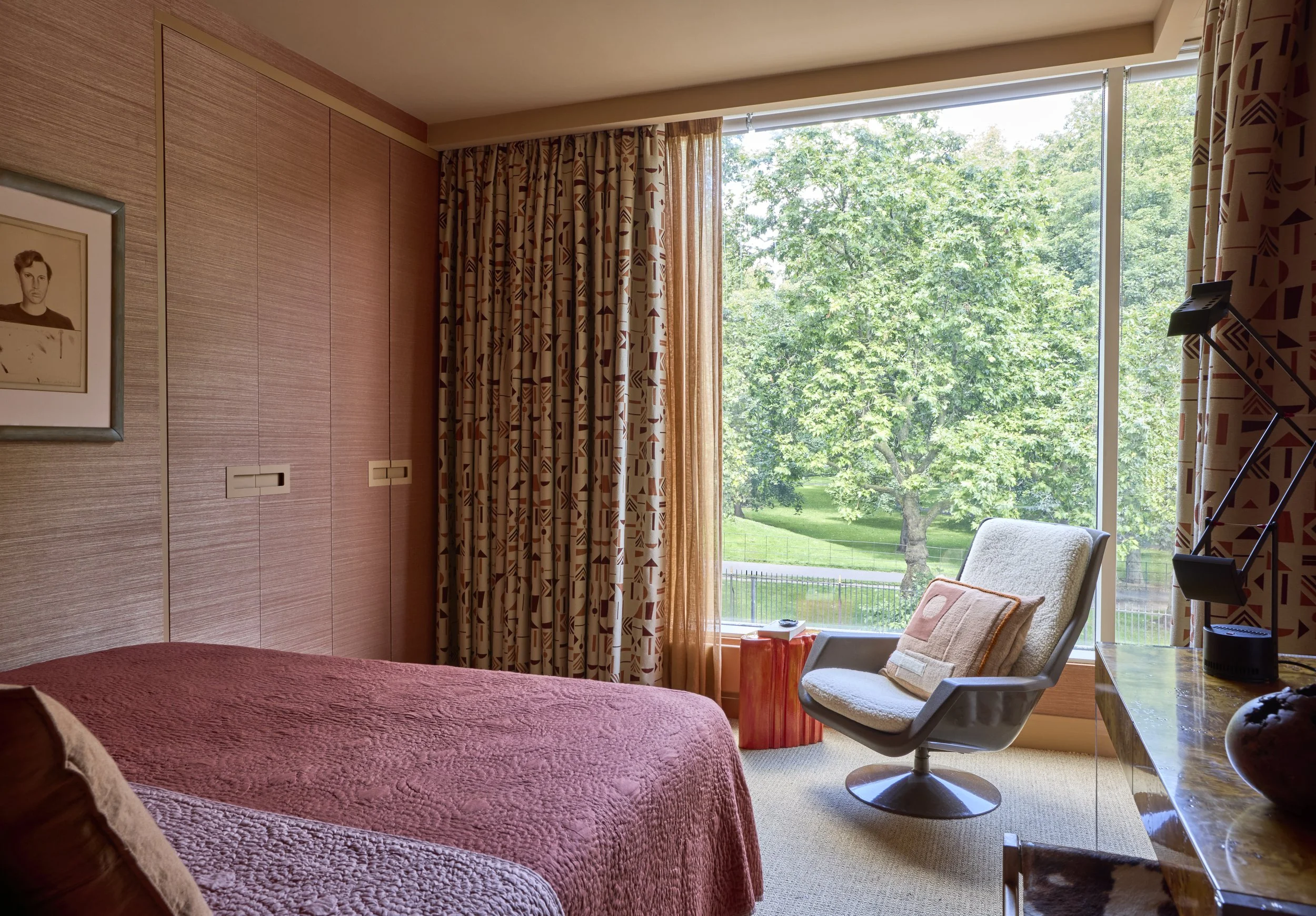 A bedroom with a large window showing green trees outside, a bed with a pink quilt, a side table, a beige armchair with a decorative pillow, and a desk with a lamp.