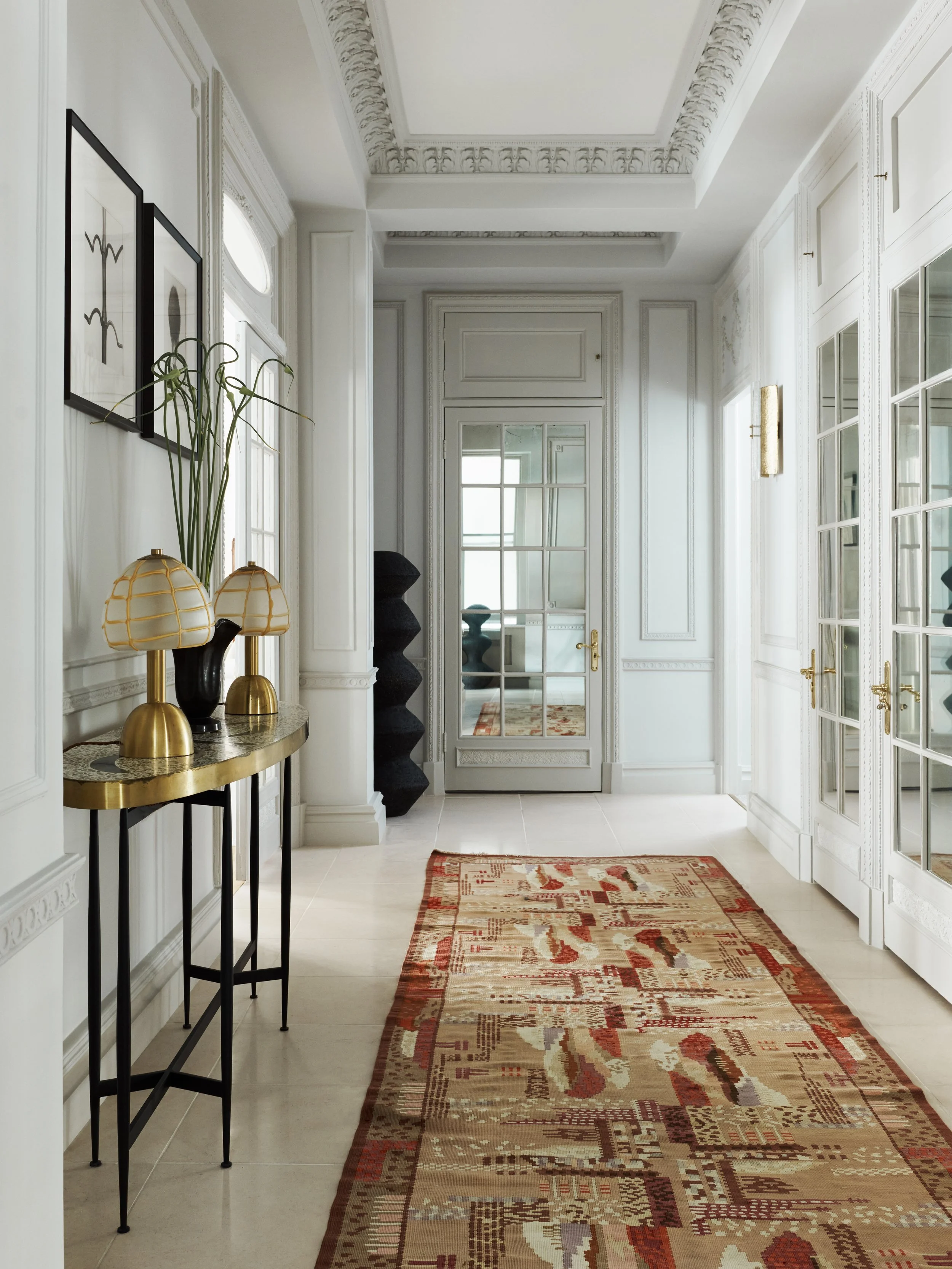 Elegant hallway with white walls, decorative molding, a Persian-style rug, a side table with gold lamps, and glass-paneled doors.