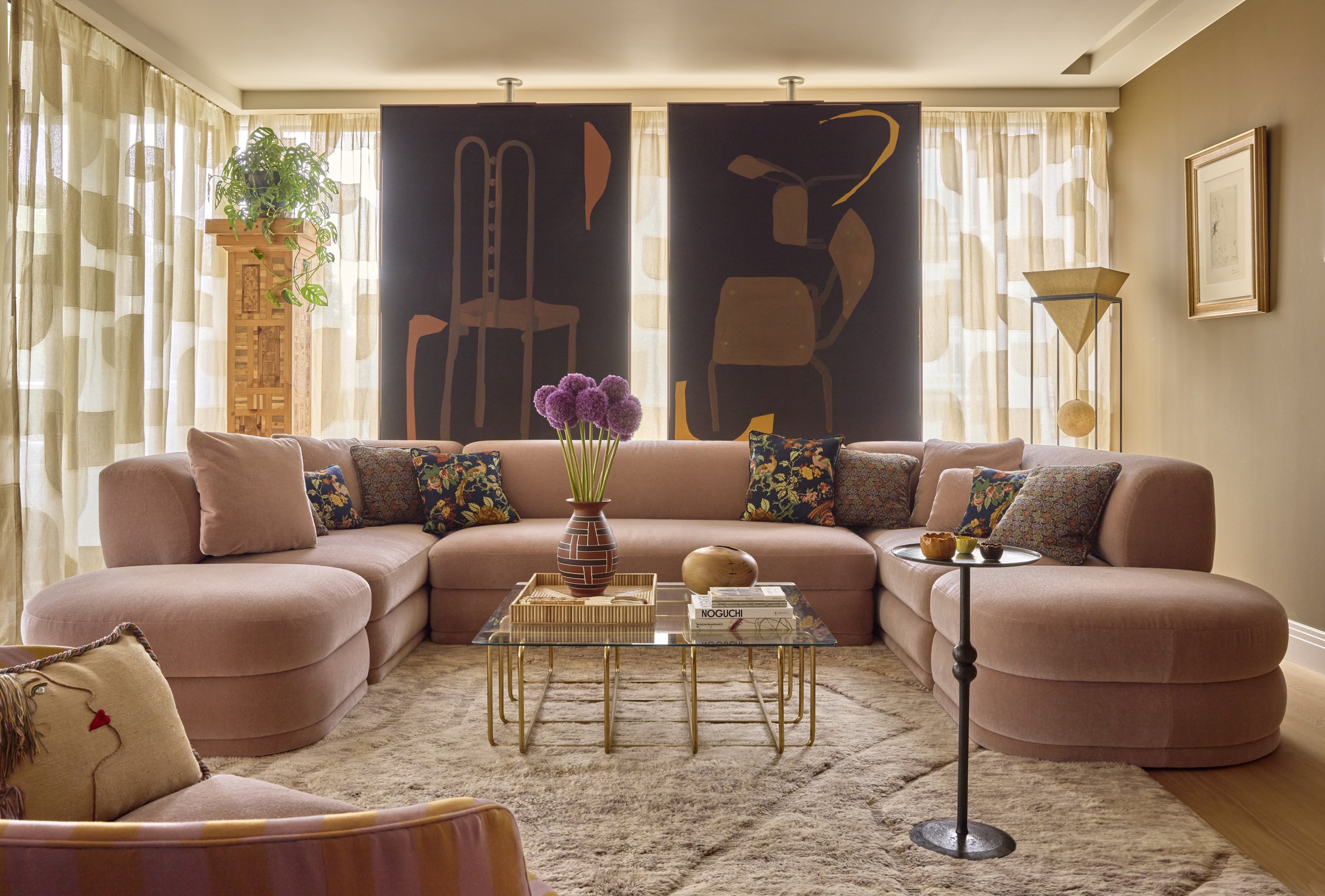 Cozy living room with a pink sectional sofa, colorful throw pillows, a glass-top coffee table with purple flower arrangement and books, window curtains with patterns, a large dark abstract art set, a tall wooden shelving unit with a plant, a side tab