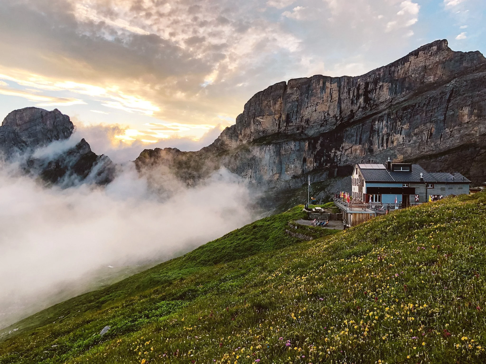 A house on a grassy hillside with colorful flowers, set against rocky mountain peaks and clouds at sunrise or sunset.