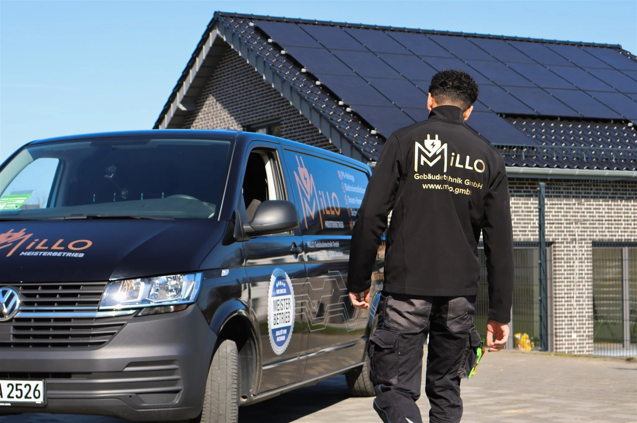 A man dressed in black work gear with the logo and website of Millo Gebäudetechnik GmbH on his back, walking next to a black Volkswagen van with the same branding, in front of a modern house with a solar panel roof, on a sunny day.
