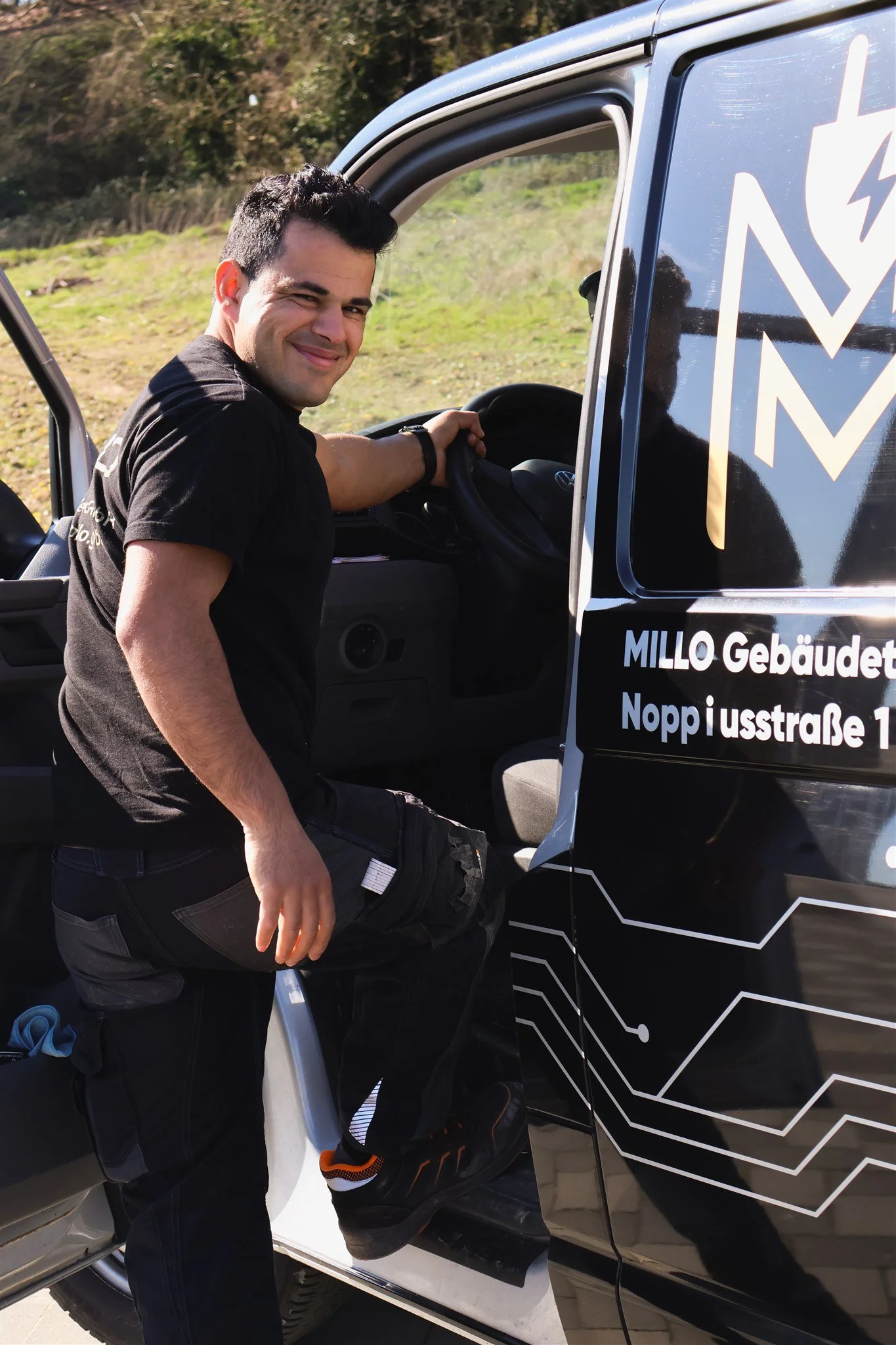 A man winking and smiling while sitting in the driver's seat of a black and white electric vehicle with the door open. The vehicle has a stylized circuit design on the side and text in German.