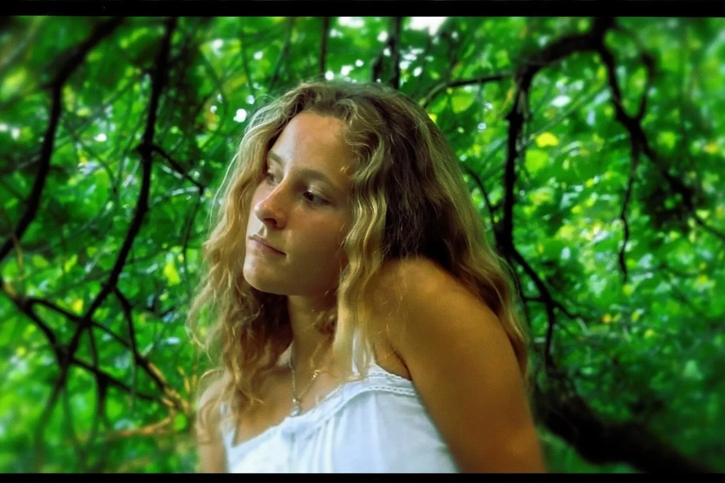 Eine junge Frau mit langen, lockigen, blonde Haare steht vor einem grünen Blätterdach im Wald.