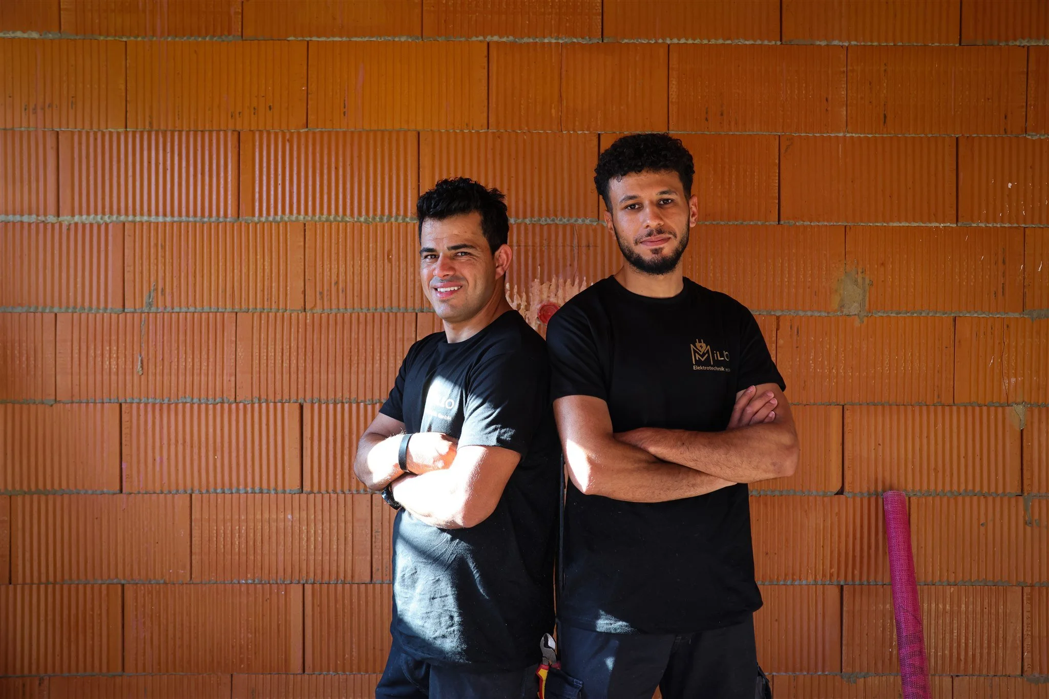 Two men are standing back to back with arms crossed in front of a brick wall. One has short black hair and a slight smile, the other has curly dark hair and a neutral expression. Both are wearing black t-shirts.