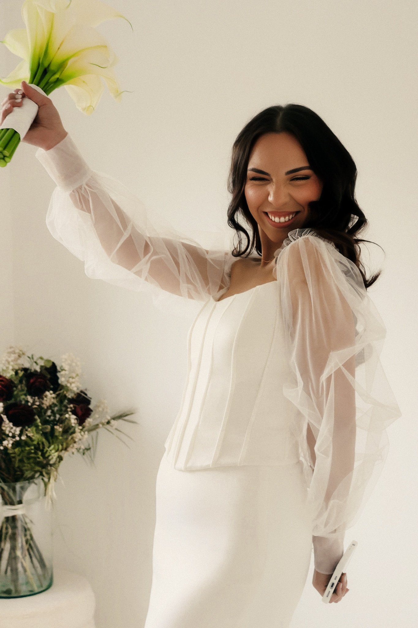 A woman with dark hair smiling and holding a bouquet of yellow lilies in one hand, while holding a smartphone in the other, standing next to a vase of flowers on a table, against a plain light-colored background.