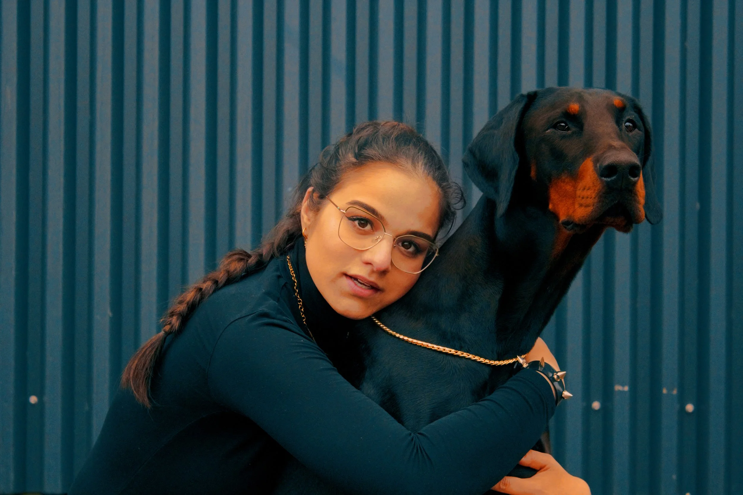 Junge Frau mit Brille und dunklem Pullover umarmt einen großen schwarzen und braunen Hund vor einer blauen Wand.