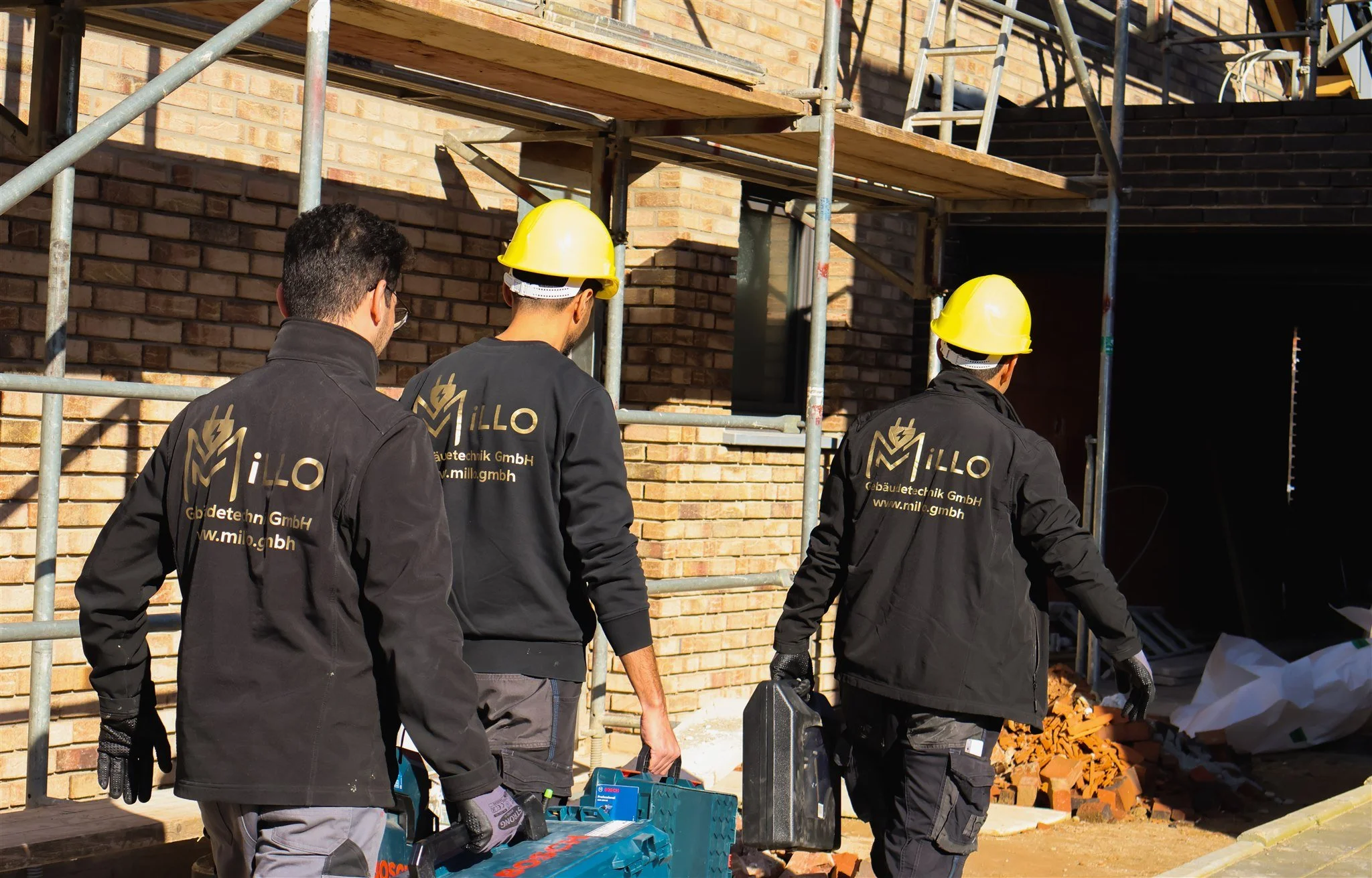 Construction workers wearing yellow hard hats and black jackets with 'Millo Bau' lettering walking near a brick building under construction.