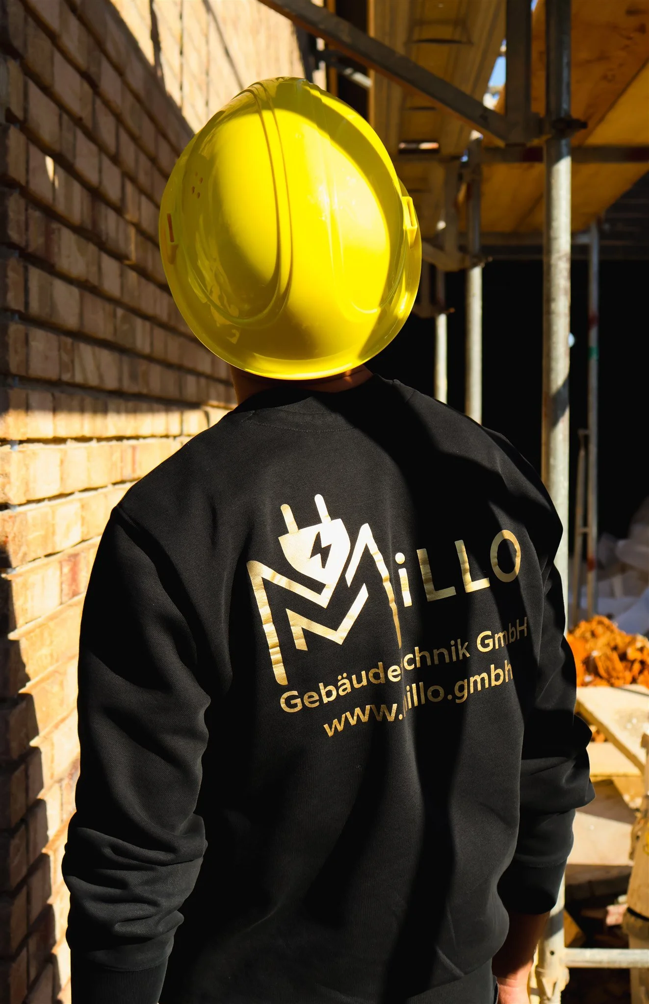 Construction worker wearing a yellow safety helmet and a black jacket with company branding, standing at a construction site with brick walls and scaffolding.