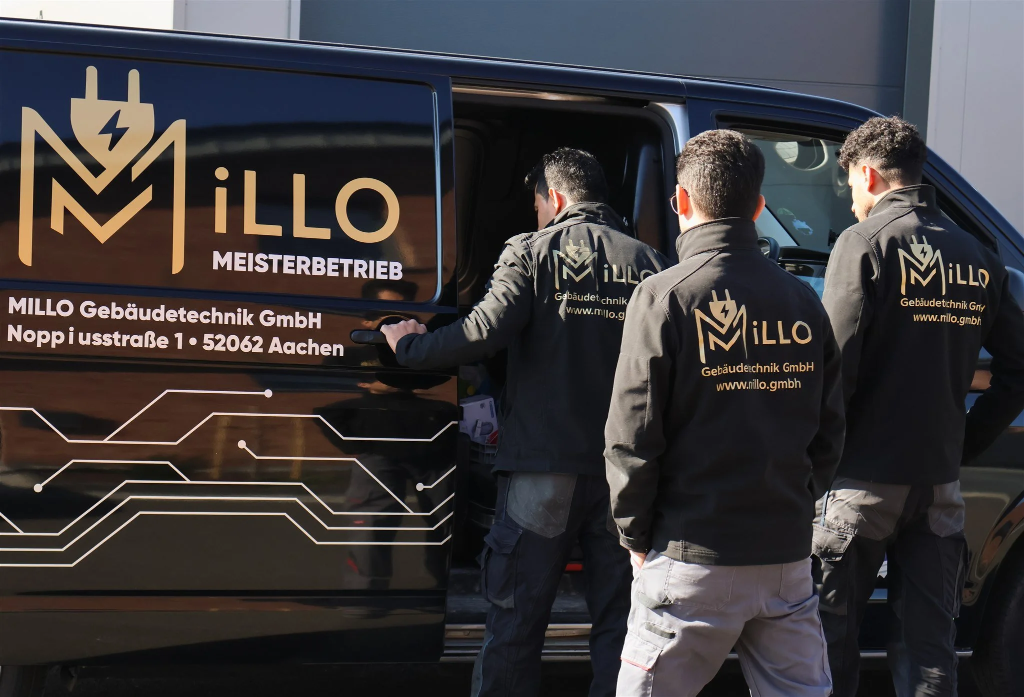 Three men wearing jackets with the logo and website of MILLO Gebäudetechnik GmbH are standing near a black van, possibly discussing something. The van has company branding and contact information on its side.