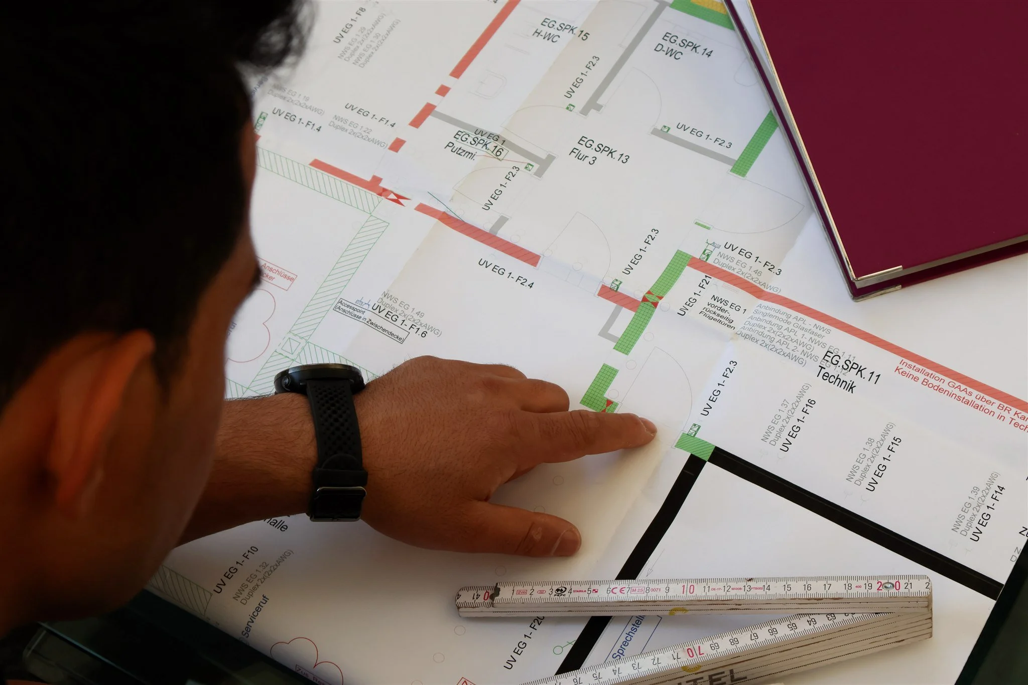 A person is examining a detailed architectural or engineering blueprint, pointing at specific sections. The blueprint includes various technical details, measurements, and color-coded areas. The person is wearing a black wristwatch and is seated at a