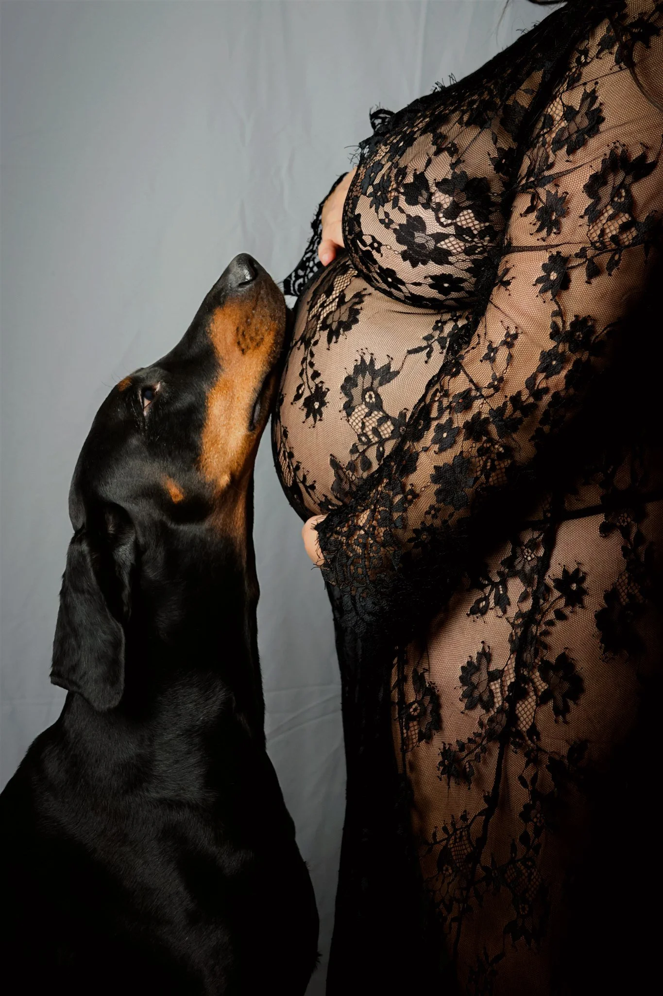 A person wearing a black lace dress with floral patterns, standing next to a black and tan Doberman dog, with the dog leaning its head against the person's chest.