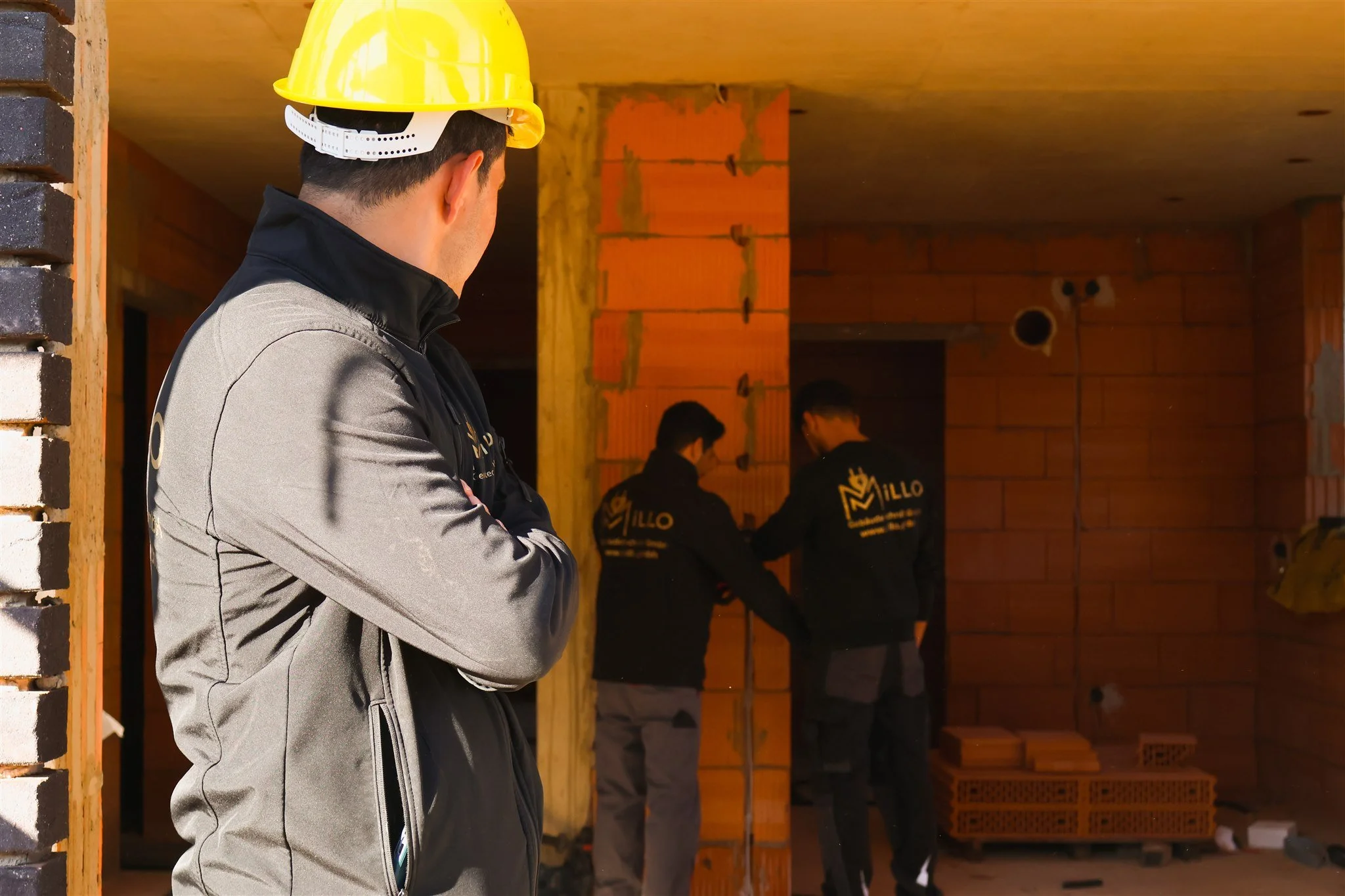 A man wearing a yellow construction helmet and a gray jacket, facing away, stands outdoors near a construction site with exposed brick walls. Two workers in black jackets with yellow logos are working in the background.