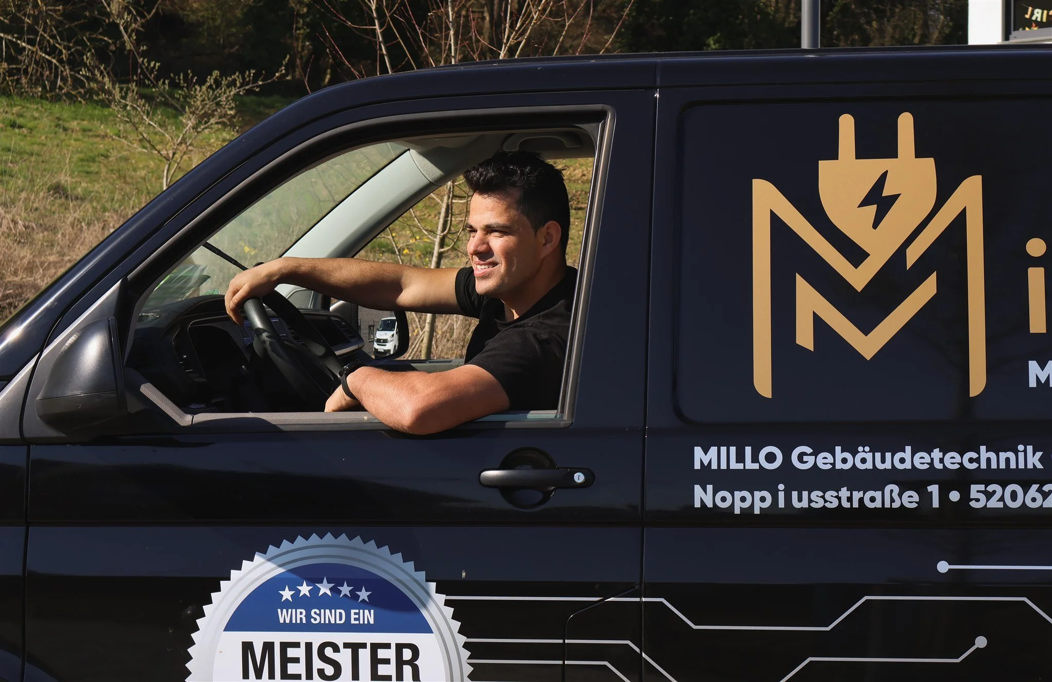 A man sitting in the driver's seat of a black company van, smiling, with promotional text and logos on the side of the van.