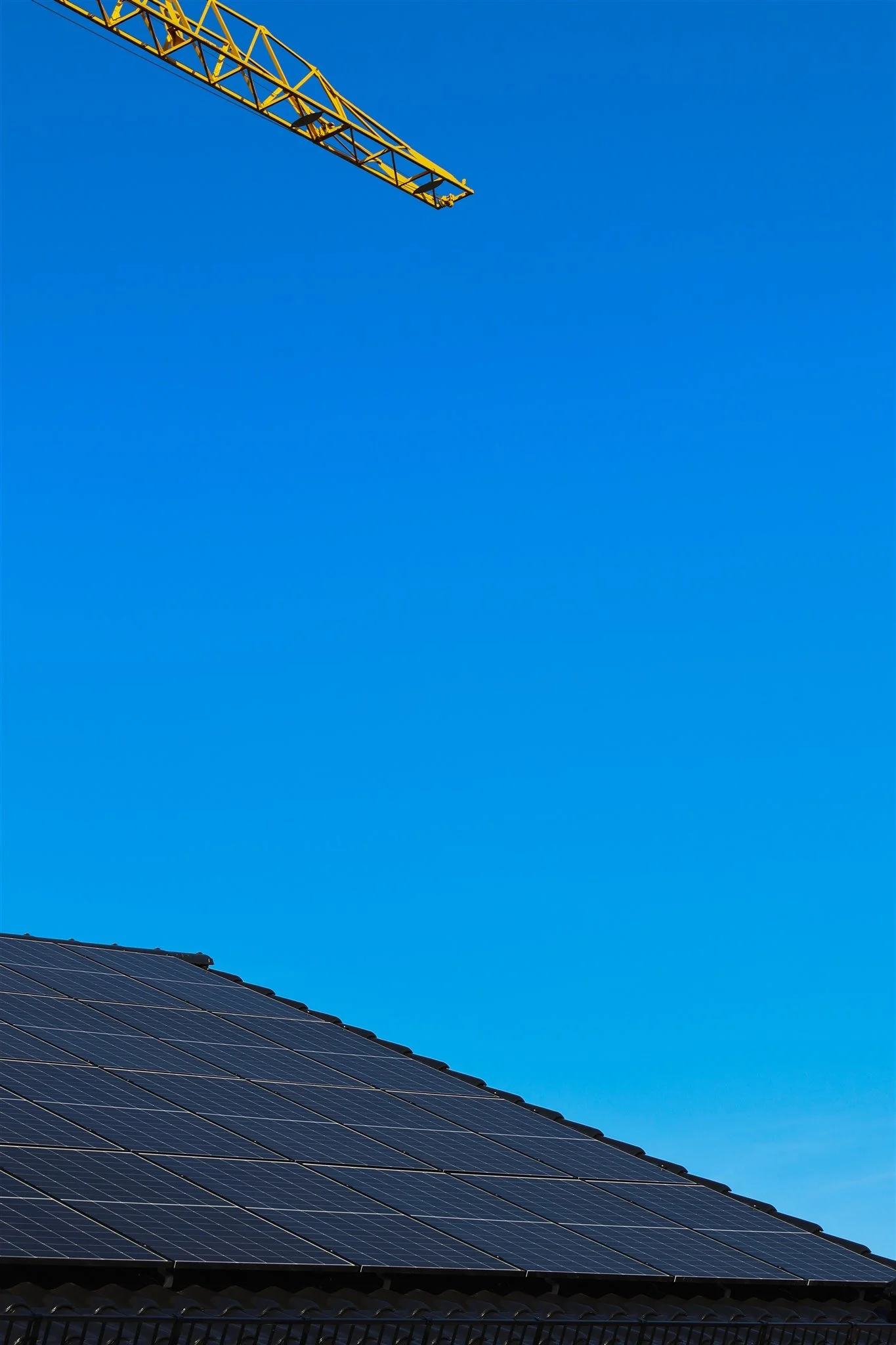 A section of solar panels on a roof beneath a bright blue sky, with a part of a yellow construction crane visible in the upper left corner.