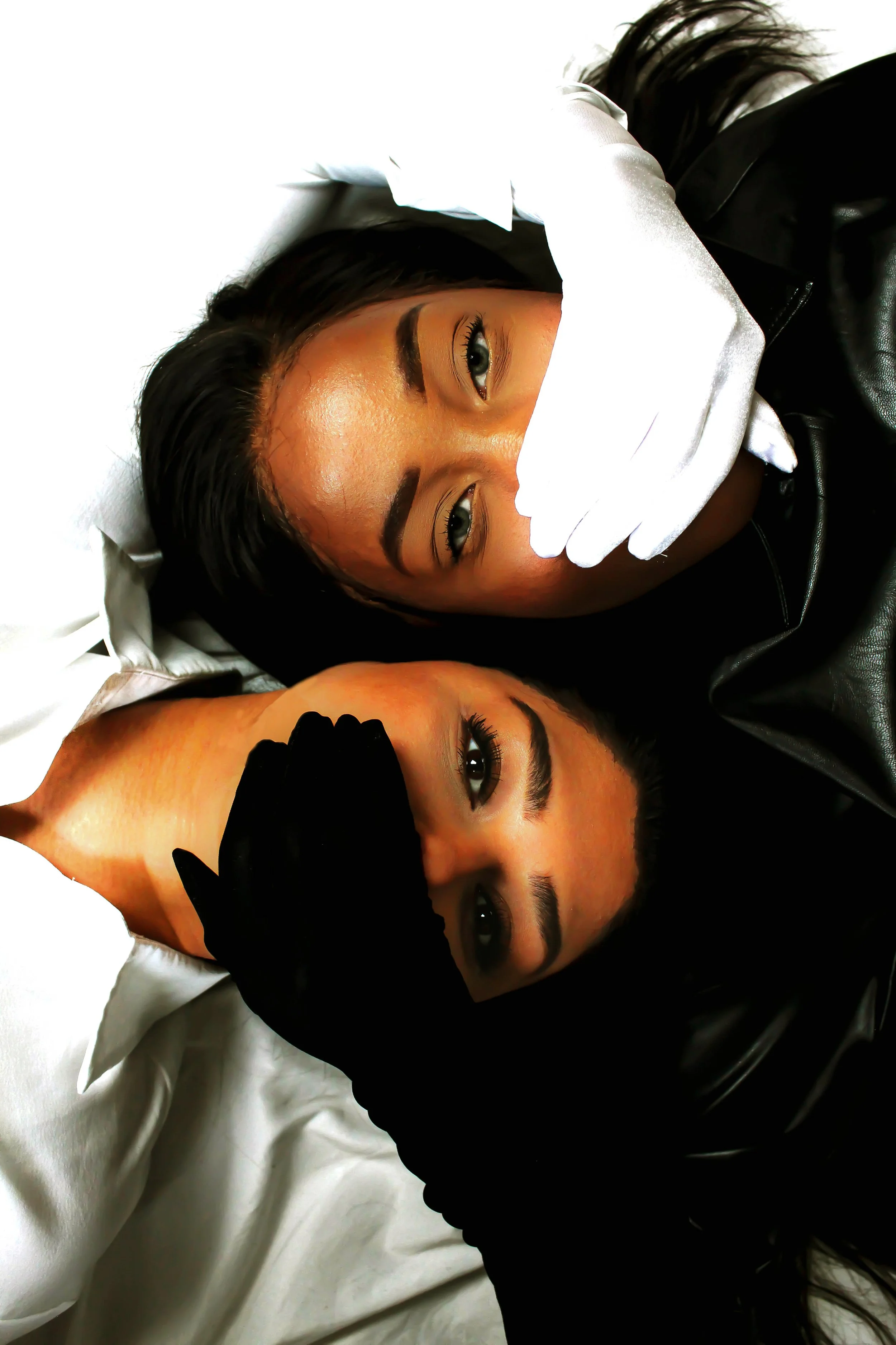 Zwei Frauen liegen nebeneinander auf einem Bett, beide schauen in die Kamera. Eine trägt schwarze Handschuhe und eine Lederjacke, die andere weiße Handschuhe und eine weiße Bluse.