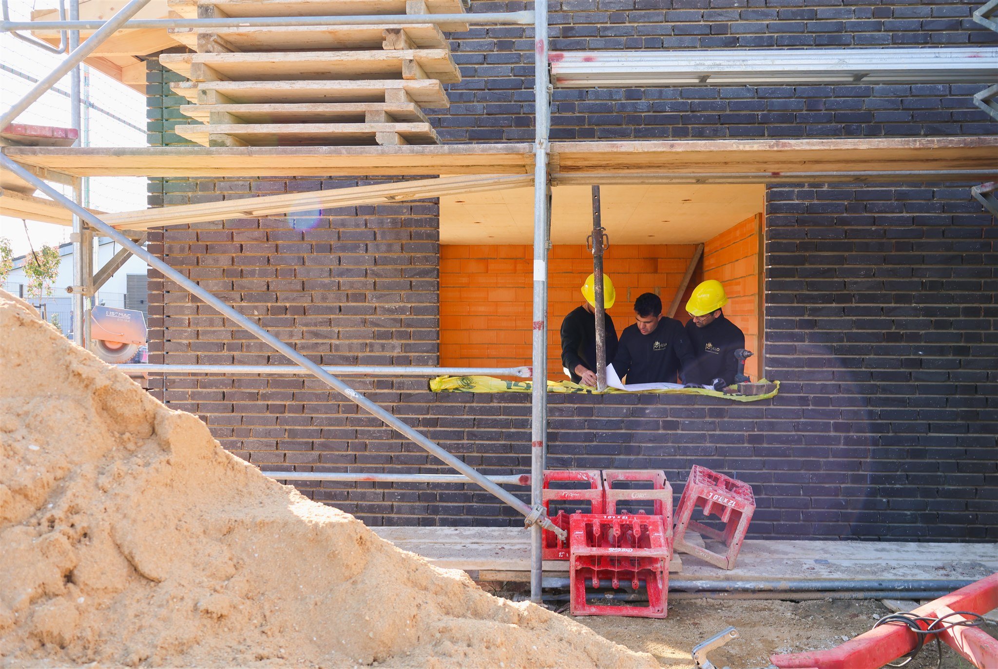 Construction workers wearing yellow safety helmets working on a building under construction, with scaffolding and brick walls, and orange brick interior visible.