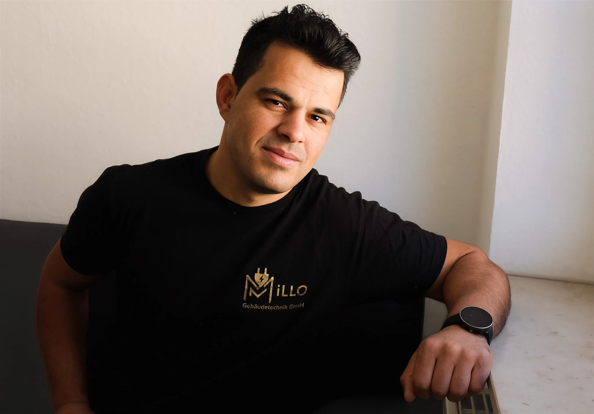 A young man with dark hair, wearing a black T-shirt with gold text and logo, sitting at a white table, wearing a smartwatch on his left wrist, and smiling slightly in front of a plain light-colored wall.