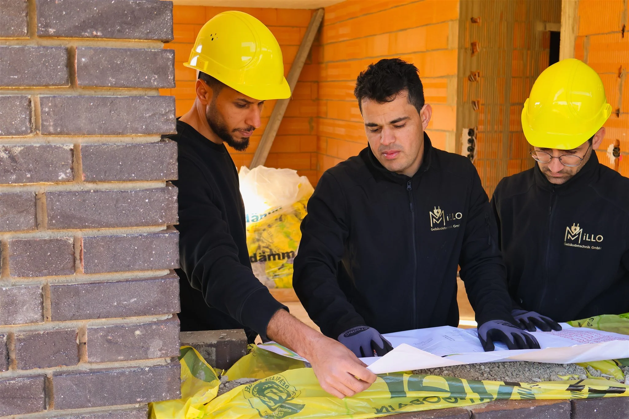 Three construction workers wearing yellow safety helmets are gathered around a table with plans or blueprints at a construction site with an exposed brick wall and unfinished interior.