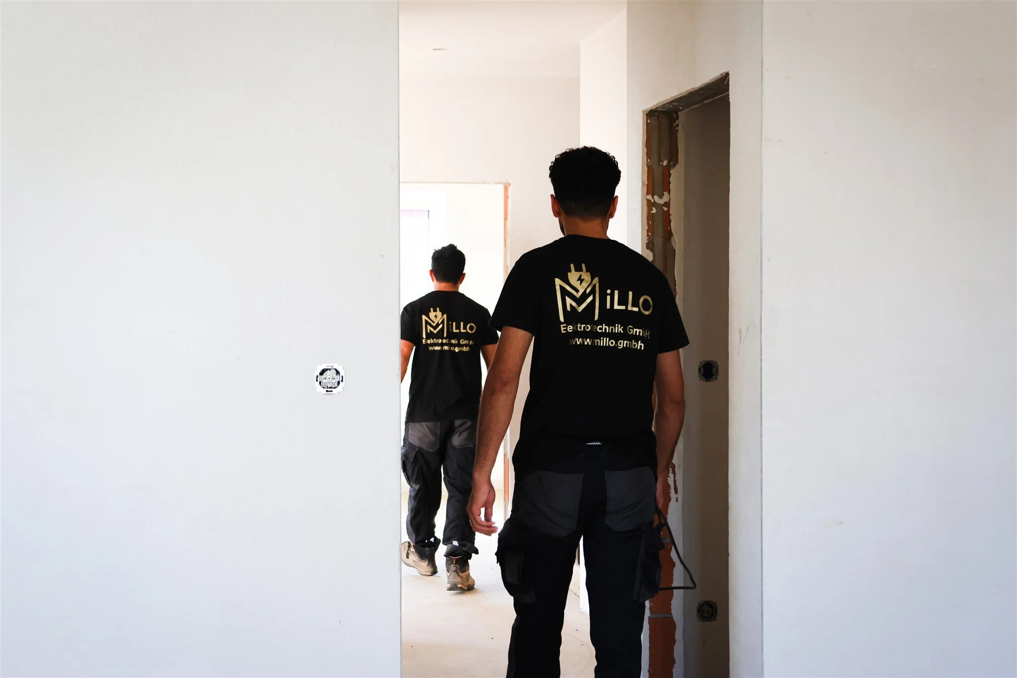 Two workers in black shirts with a logo and website walking through an unfinished room with white walls and exposed door frames.