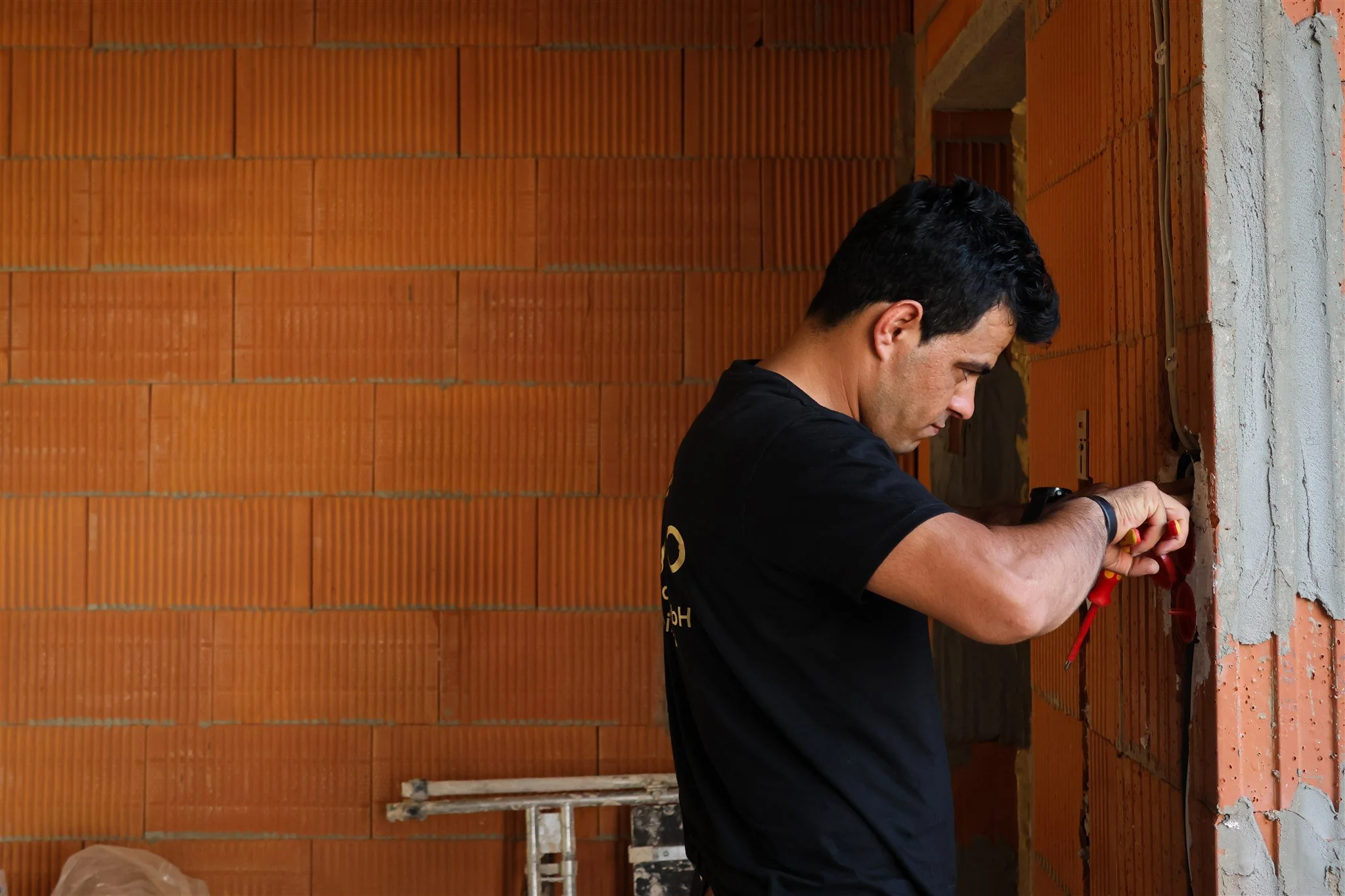 Man working on electrical wiring inside a building with exposed brick wall.
