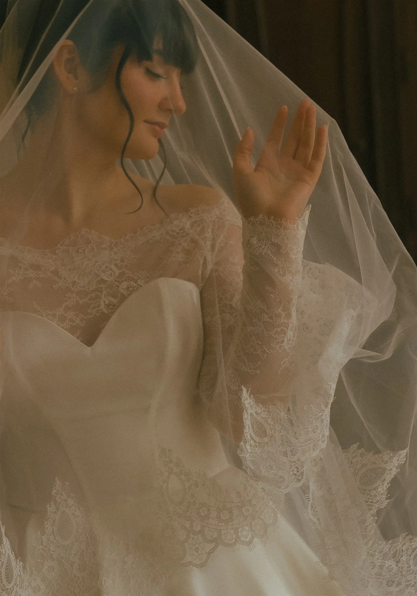 A bride wearing a lace wedding dress and veil, with dark hair in an updo, touching her veil, on a dimly lit background. cinematic wedding photography. Eine Braut in Hochzeitskleid und Schleier. Film Hochzeitsfotografie, Hochzeitsfotografin Aachen