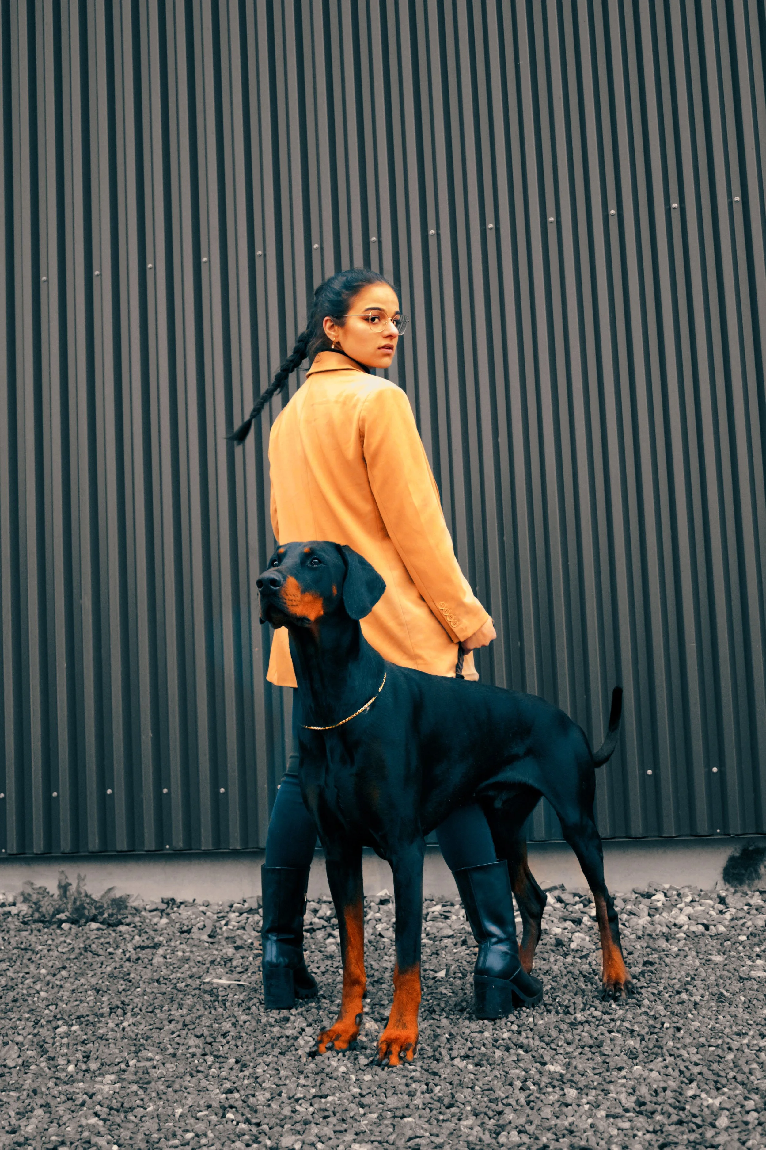 Junge Frau mit Brille in orange Jacke hält einen schwarzen und braunen Dogge an der Leine vor einer dunklen, gestreiften Wand.