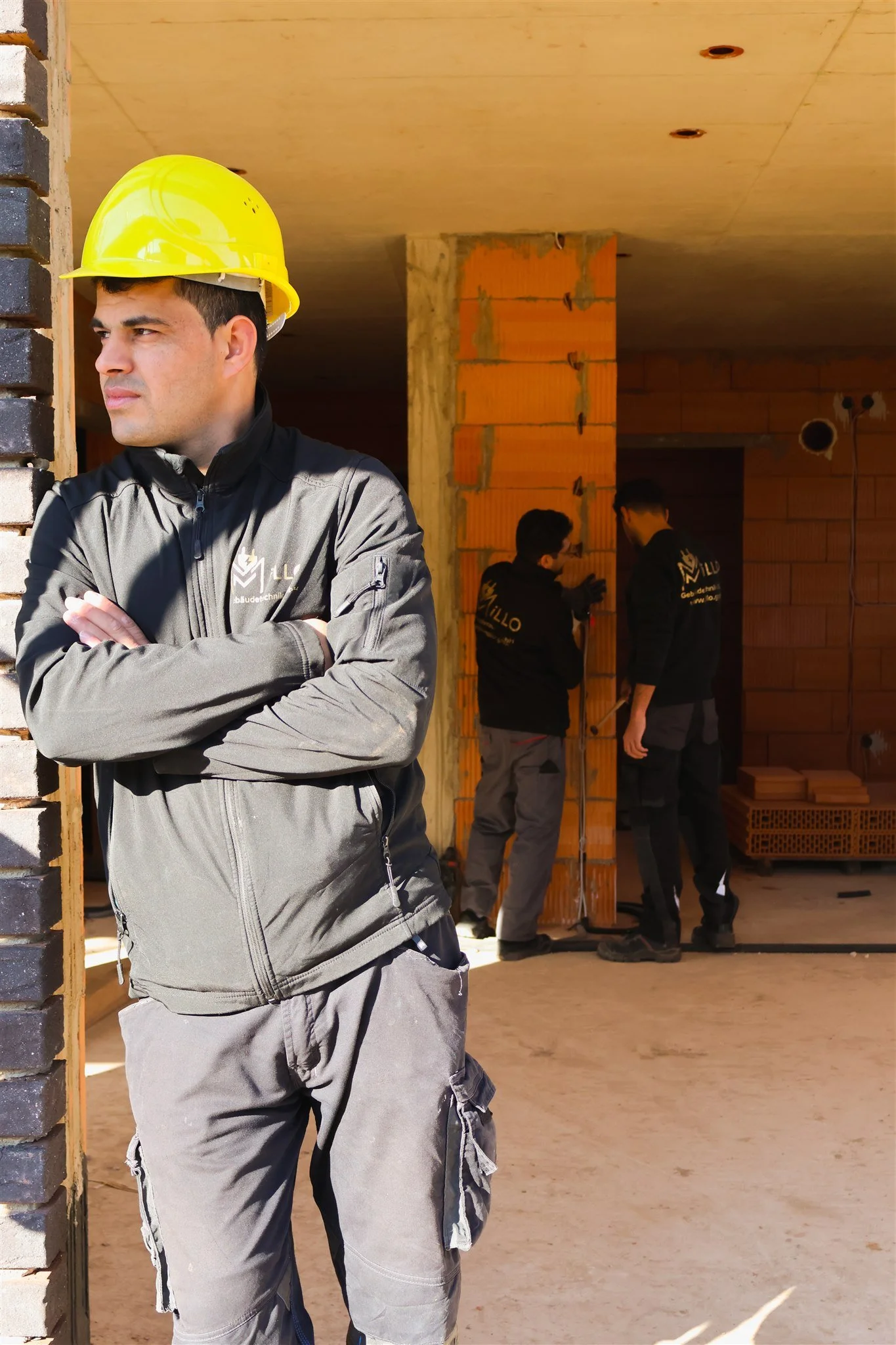 Construction workers at a building site, with a man wearing a yellow safety helmet and black jacket in the foreground, and three men in black jackets talking in the background inside an unfinished structure.