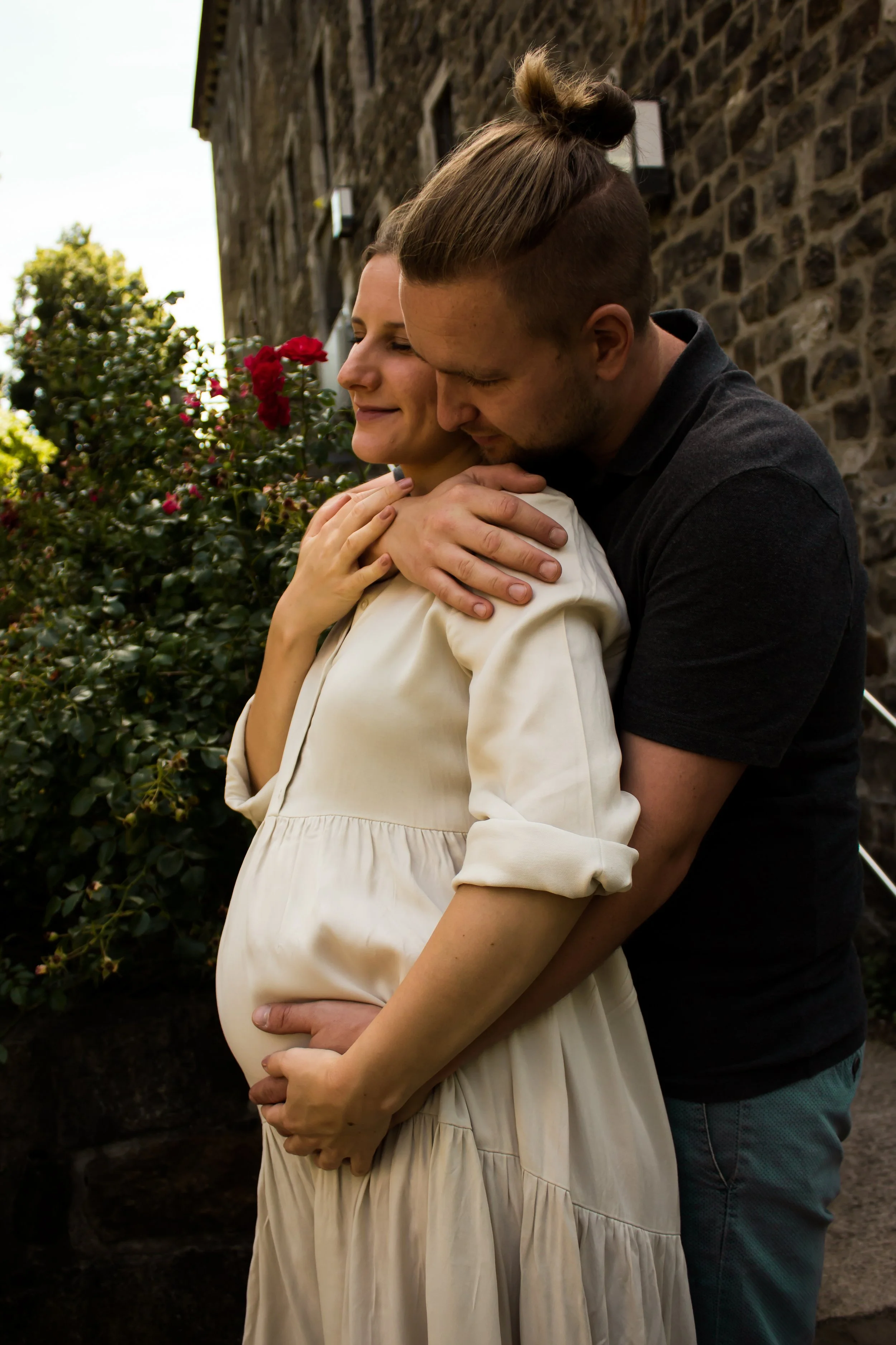 Ein Paar umarmt sich liebevoll vor einem Haus mit Steinmauer, die schwangere Frau trägt ein weißes Kleid, der Mann trägt ein schwarzes T-Shirt.