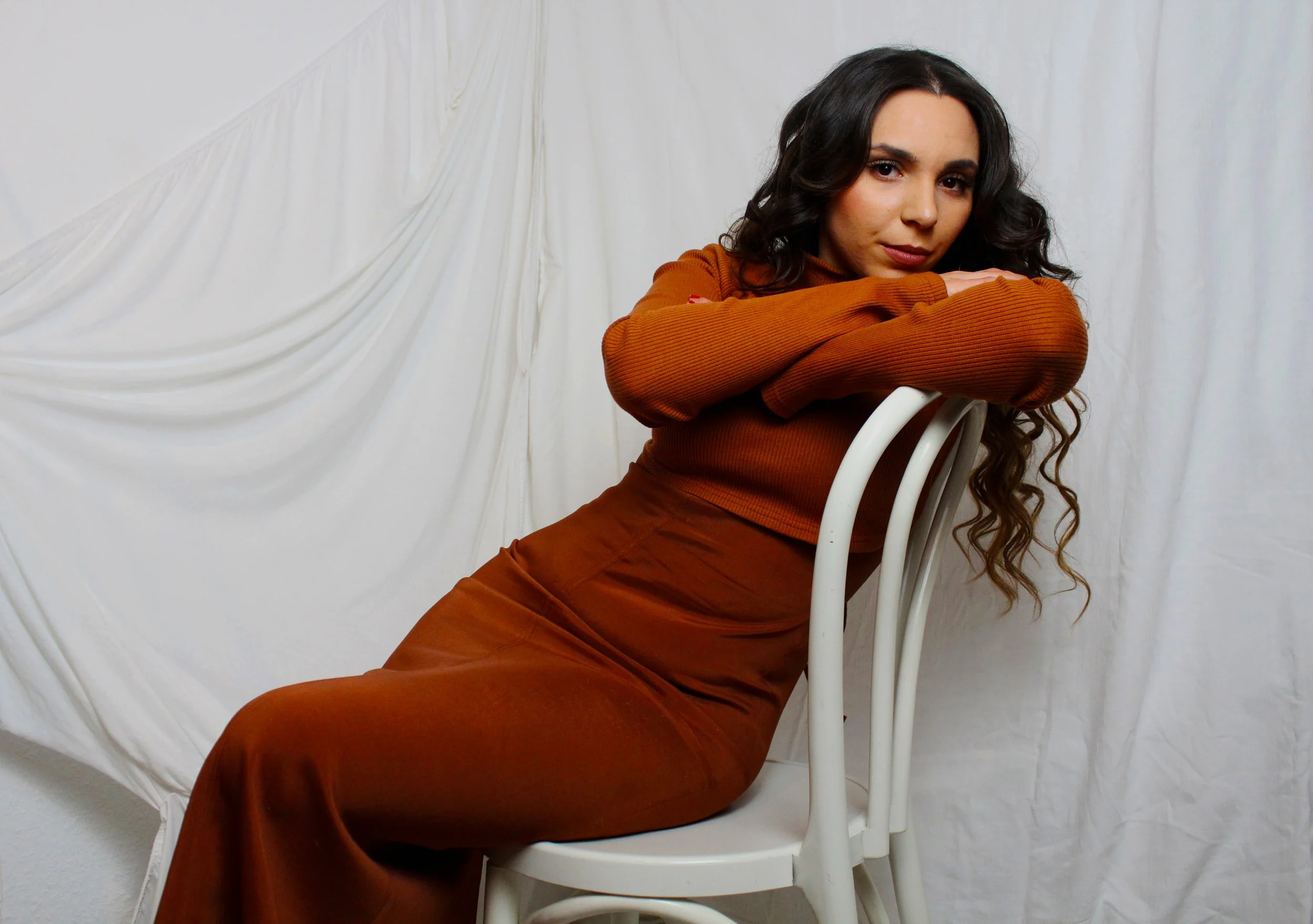 Frau mit langen, dunklen, gewellten Haaren lehnt auf einem weißen Stuhl, trägt einen braunen Pullover und einen Braunen Rock, vor einem weißen Stoffhintergrund.