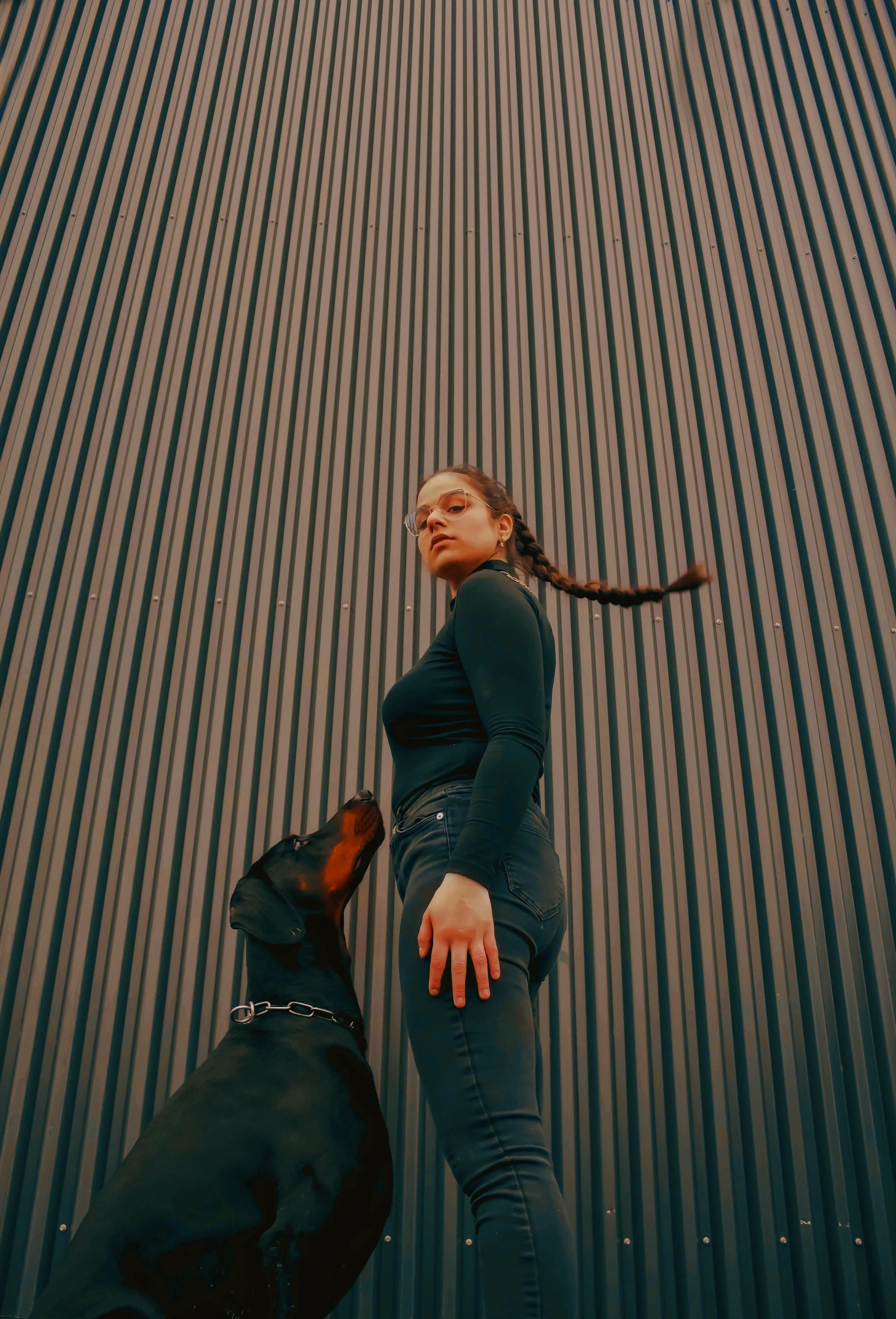 Junge Frau mit Brille steht vor einer Wand mit vertikalen Linien, ein Hund schaut zu ihr hoch.