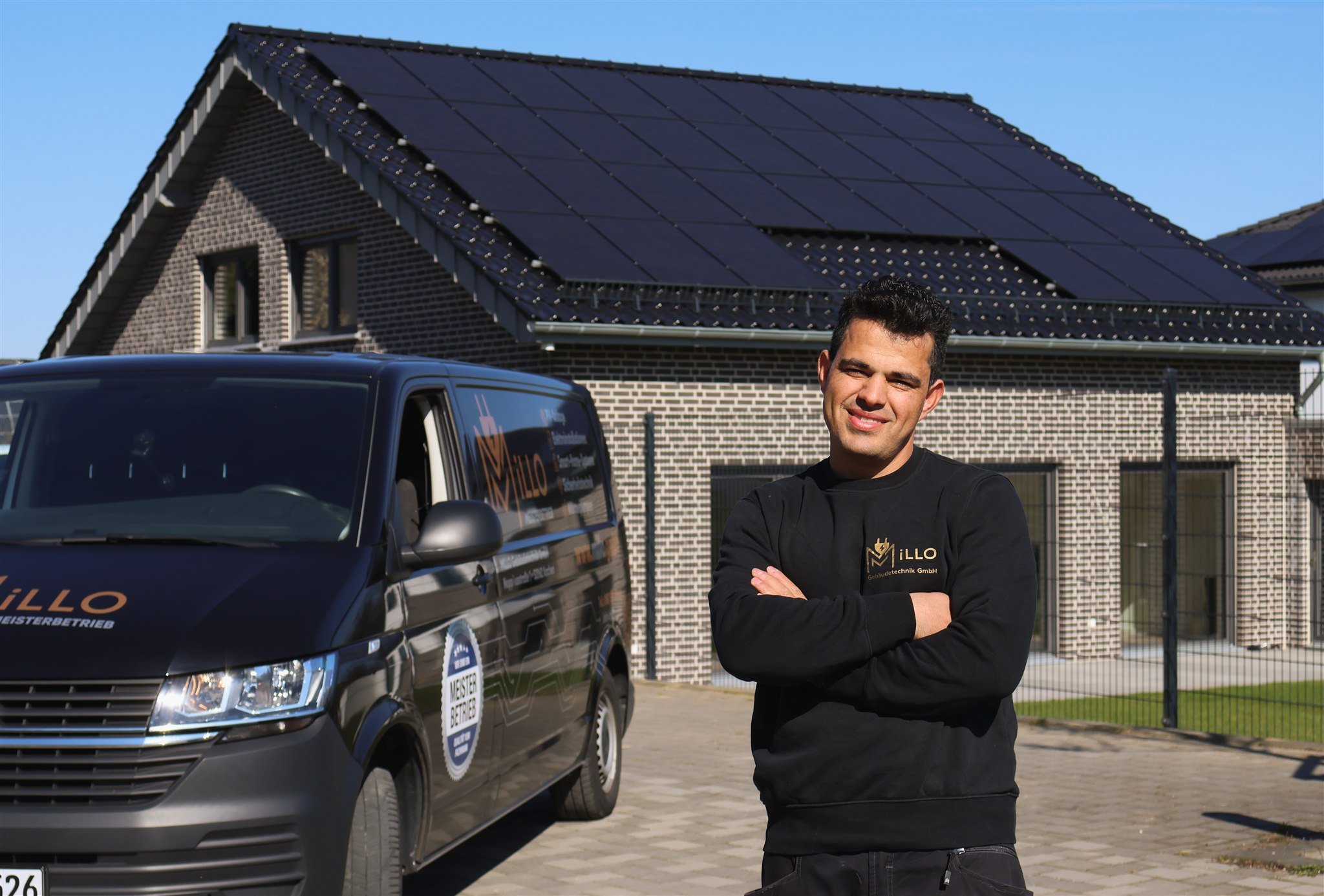 A man standing with crossed arms in front of a black van with solar panels on its roof, parked outside a house with a brick exterior and a roof also covered with solar panels on a clear day.