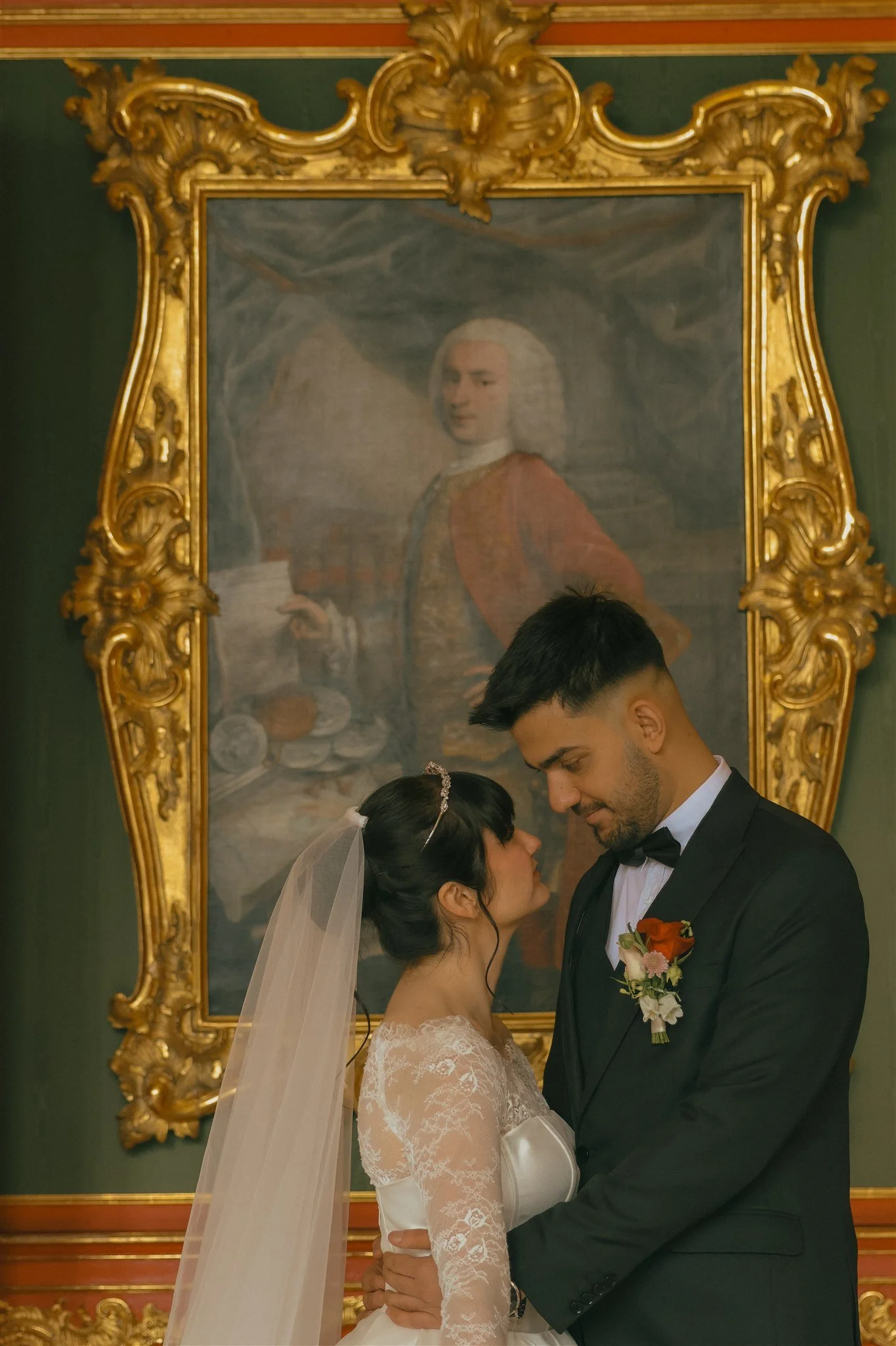 A bride and groom are standing close to each other, about to kiss. The bride is wearing a white lace wedding dress and a veil, and the groom is dressed in a black tuxedo with a bow tie and boutonniere. They are in an opulent room with a large, ornate gold-framed portrait of a woman in historical dress hanging behind them.