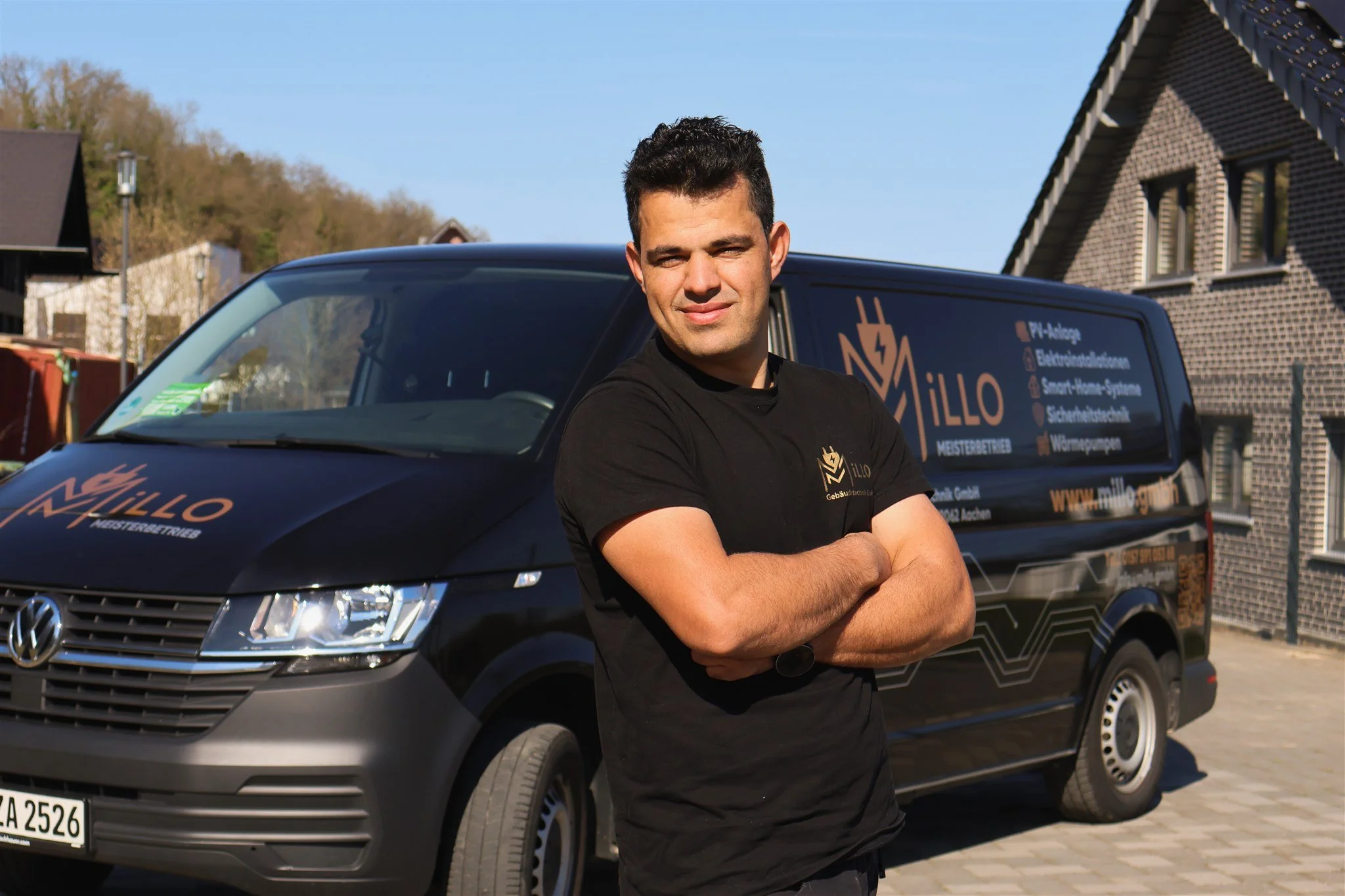 A man with short dark hair and a black T-shirt standing with arms crossed in front of a black van with company branding, outdoors on a sunny day with a house and trees in the background.