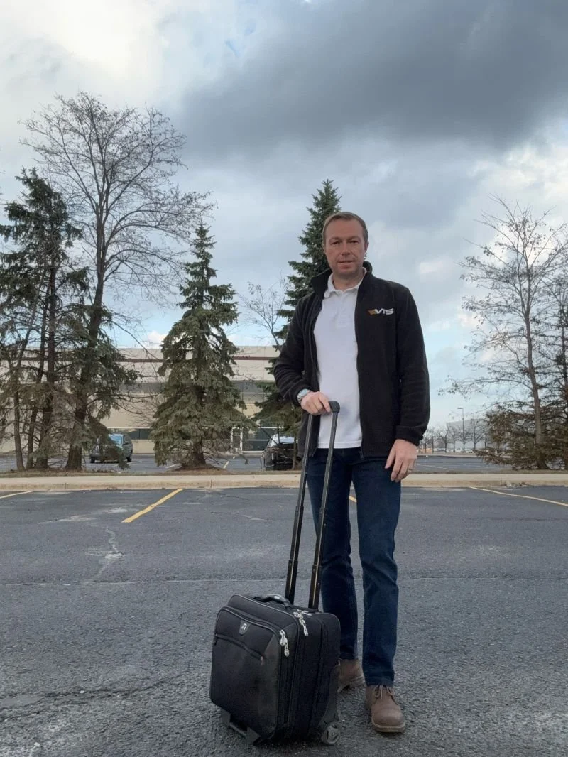 Scott Lindsay standing in an empty parking lot holding a rolling suitcase VTS Online