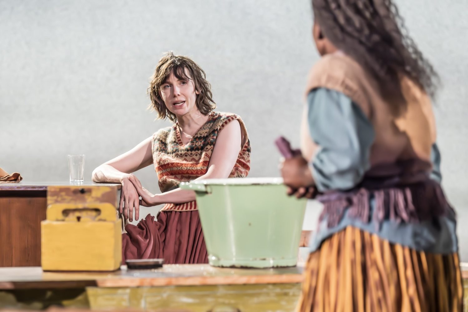 A woman with short, wavy hair, wearing a patterned sleeveless top, sits at a countertop looking frustrated or upset, with a glass of water in front of her. In the foreground, a person with curly hair, wearing a layered vest and striped skirt, is hold