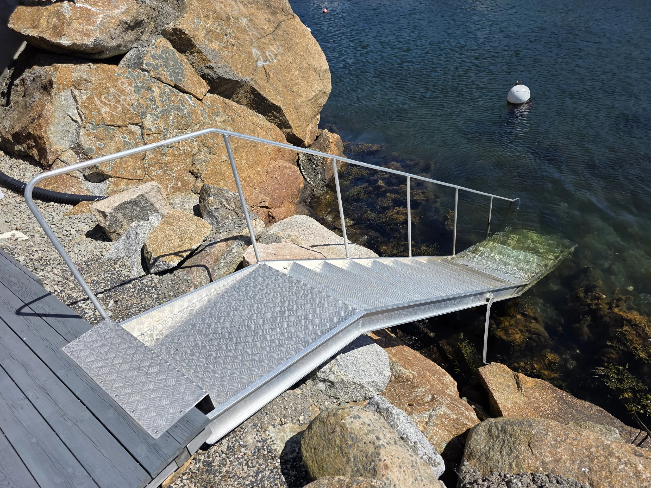 En metallramp fører ned til vannet ved en steinbelagt strand, omgitt av store steiner og hav. Det er en hvit badebøye i vannet.