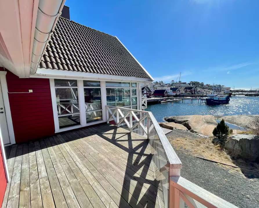 Utsikt fra terrassen på en trehus med utsikt over sjøen, båter og nabohus, med solen som reflekteres i vannet.