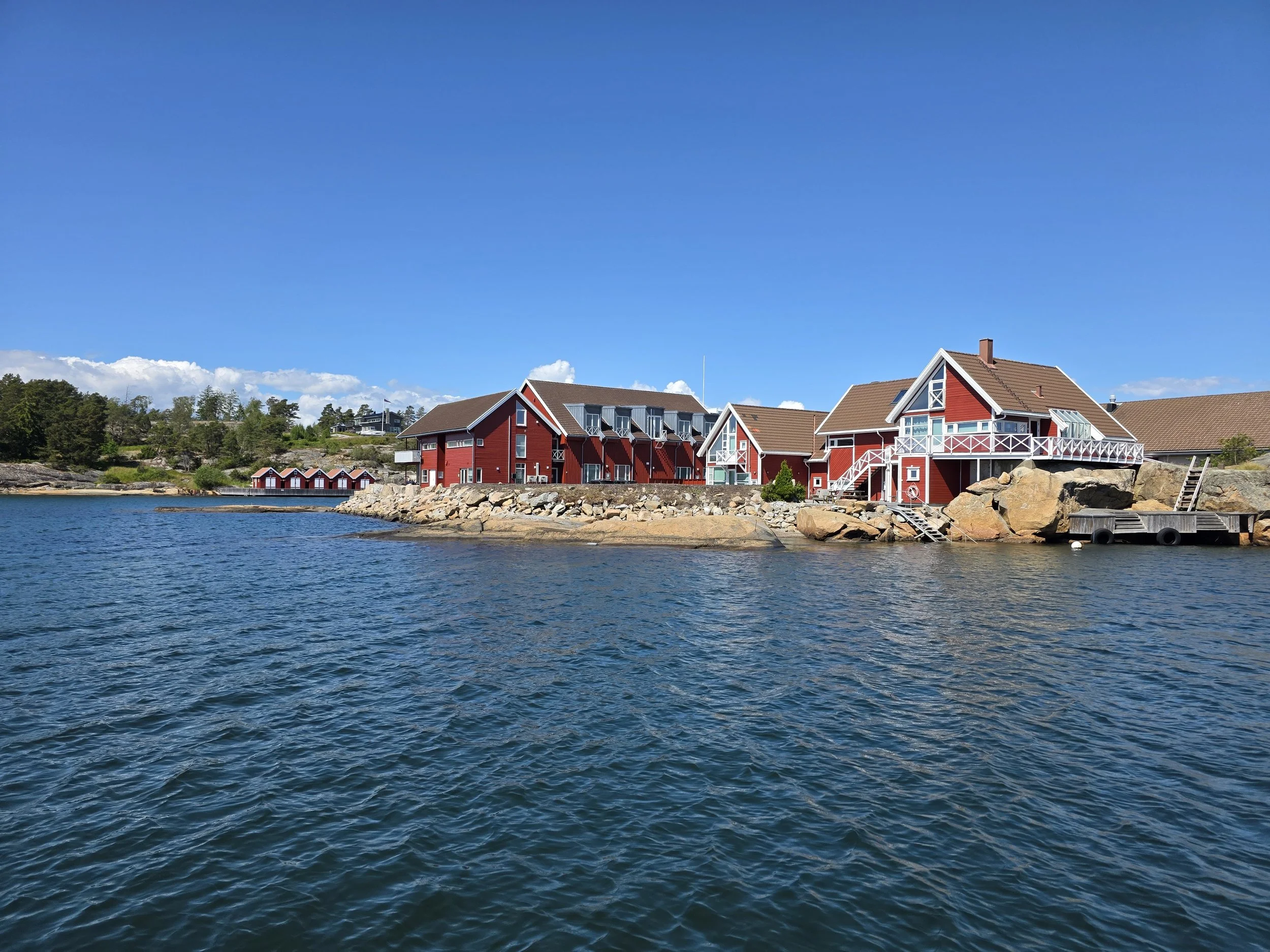 Røde trehus ved vannkanten på en klar dag med blå himmel og noen skyer.
