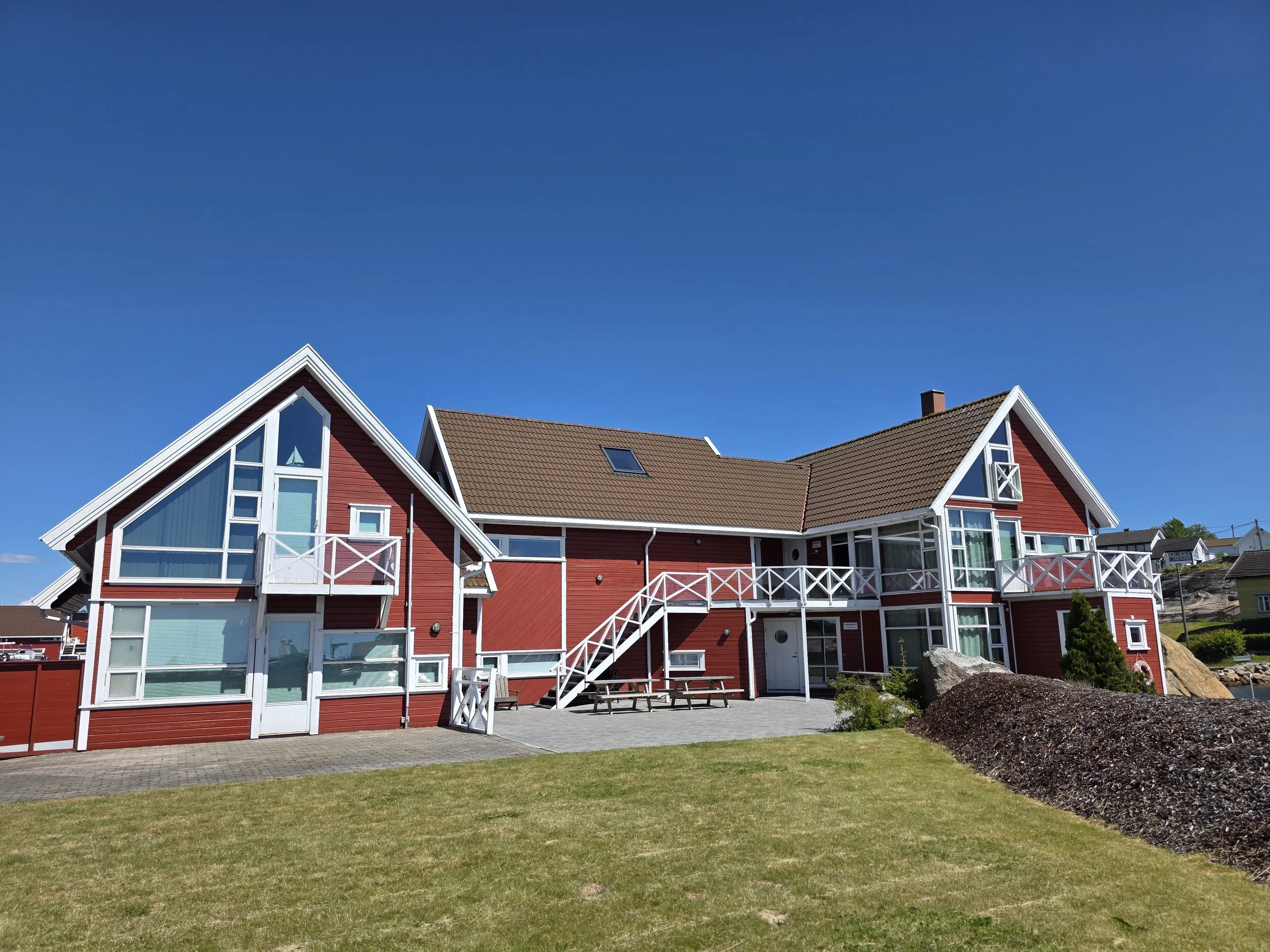 Rødt hus med hvite vindusrammer og balkonger, omgitt av grønt område under en klar blå himmel.