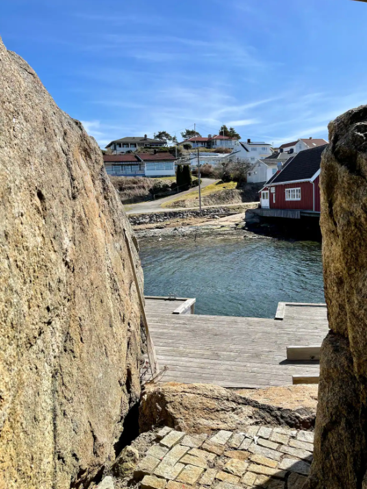 Utsikt gjennom en åpning mellom to store steiner mot en liten båtutside ved vannet, med hus i bakgrunnen på en åsside.