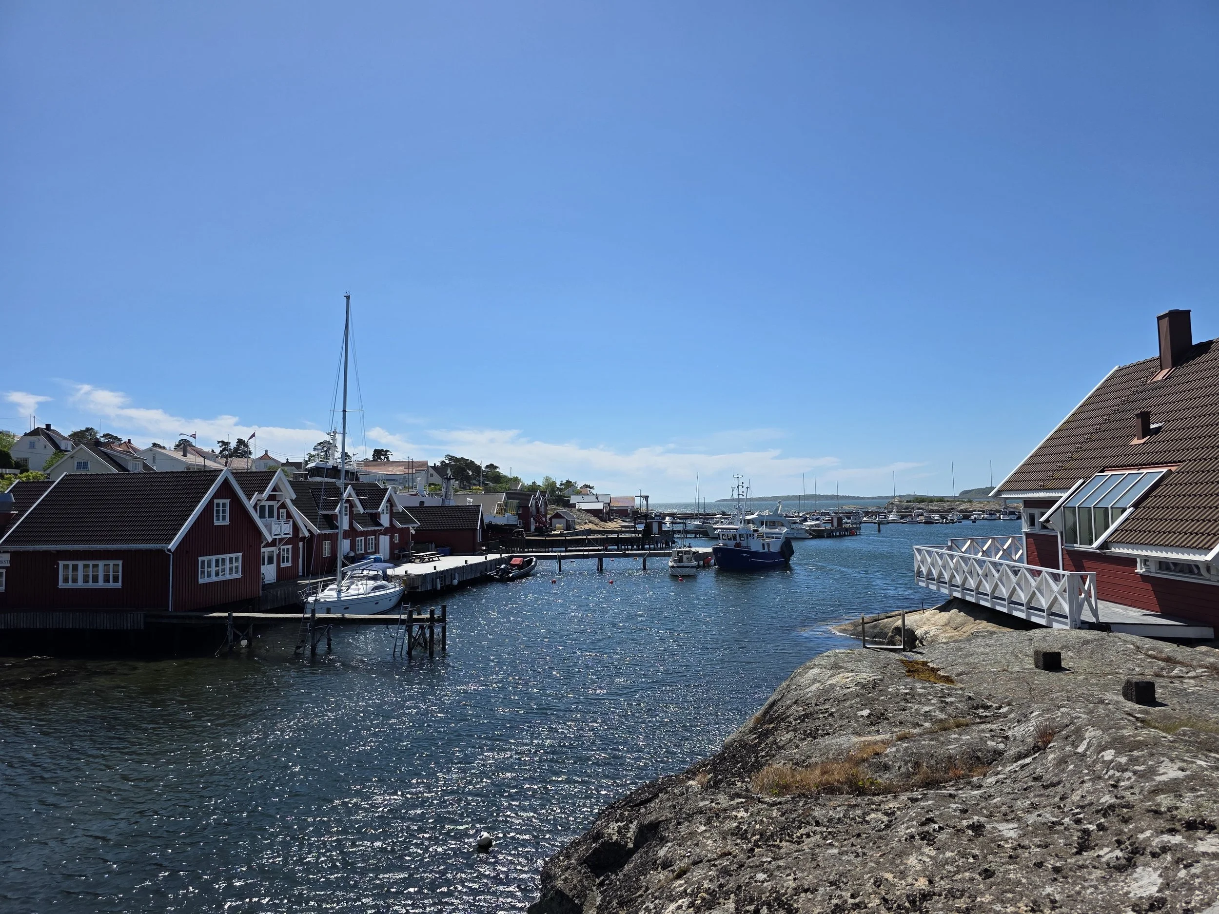 Foto av en havn med båter og røde hus, solskinn og klar blå himmel.