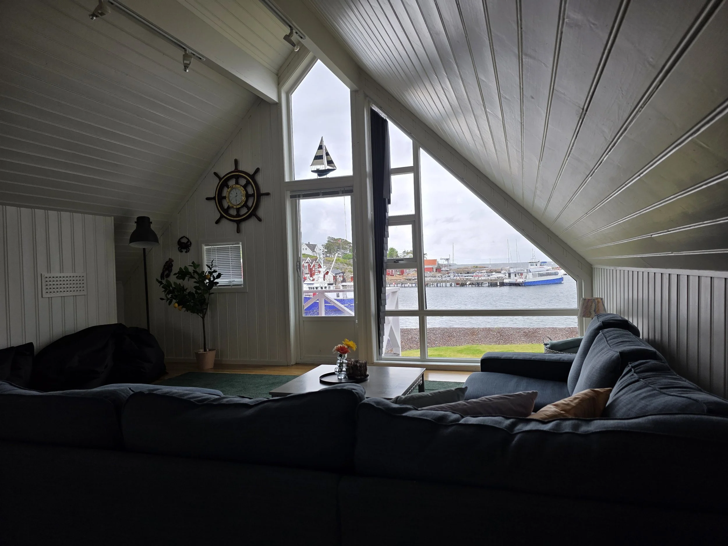 Stue med stor sofa, vindu med utsikt over havnen og båter, maritime dekorasjoner, lampe og plante.