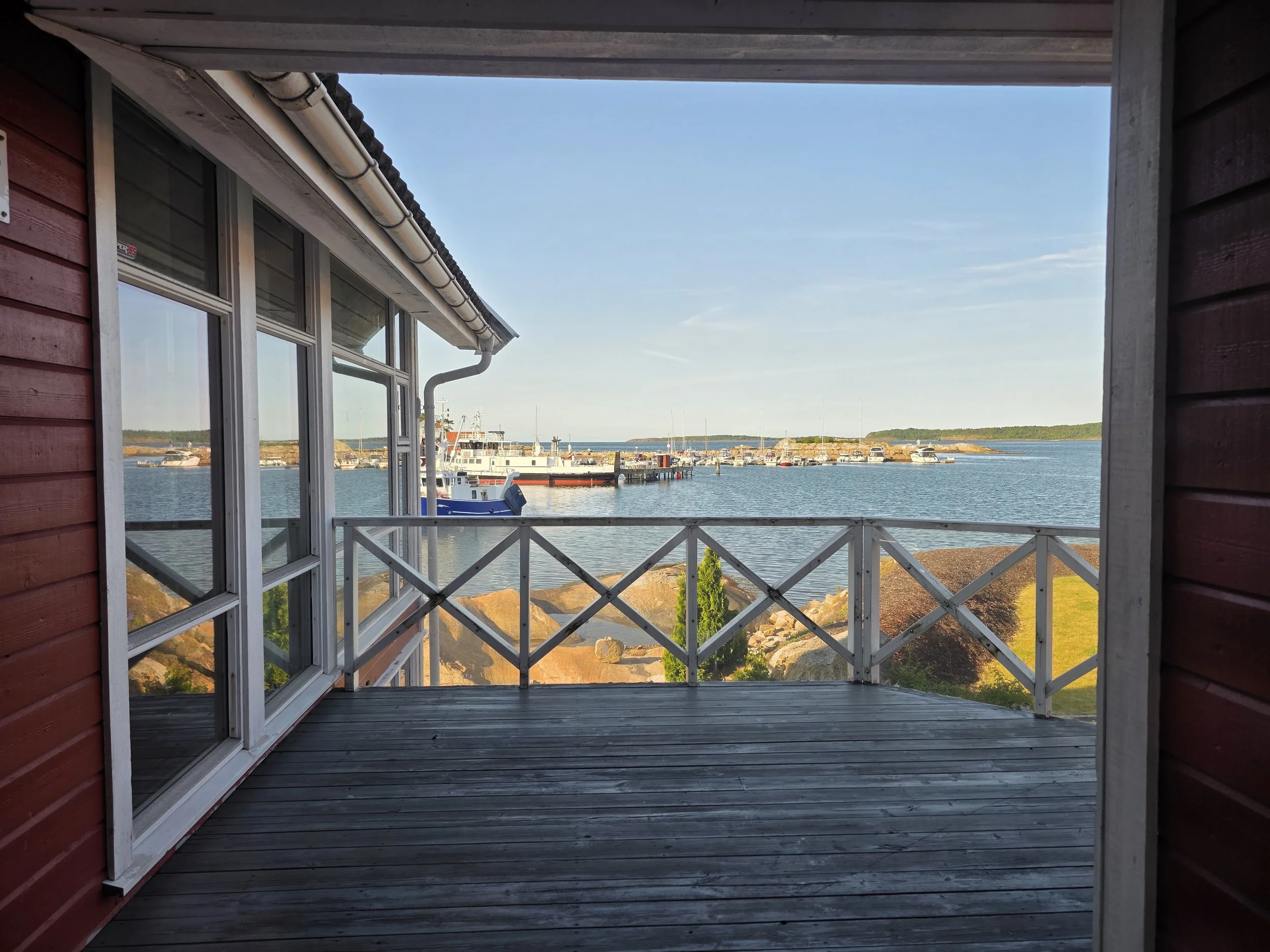 Utsikt fra en veranda over vann med båter og en liten øy i bakgrunnen, omgitt av sjø og blomstrende natur, sett fra innsiden av et hus med store vinduer og mocks.