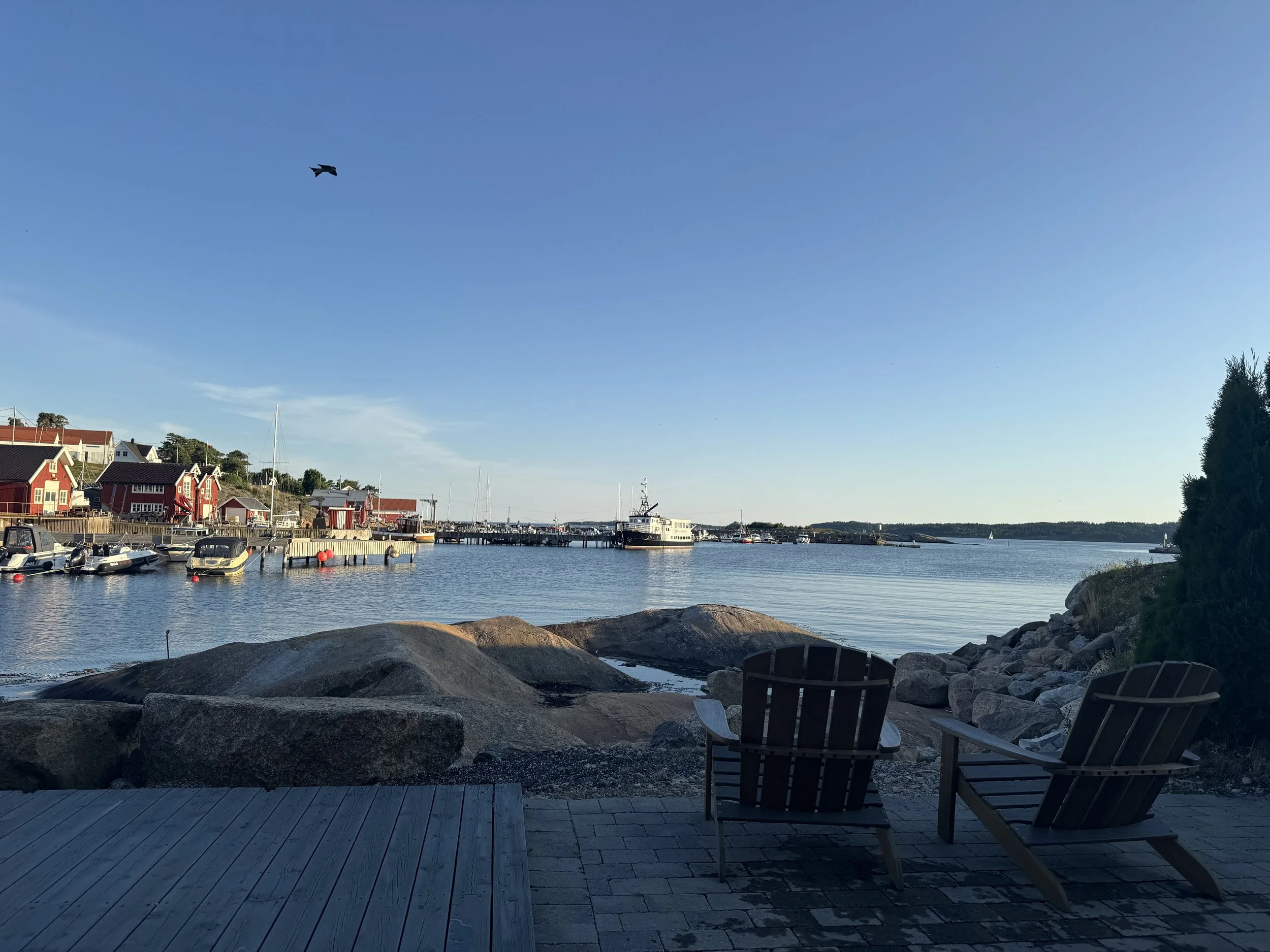 Utsikt over en havn med kai, båter, hus, og et fly som flyr i en klar blå himmel, sett fra en terrasse med to Adirondack-stoler.