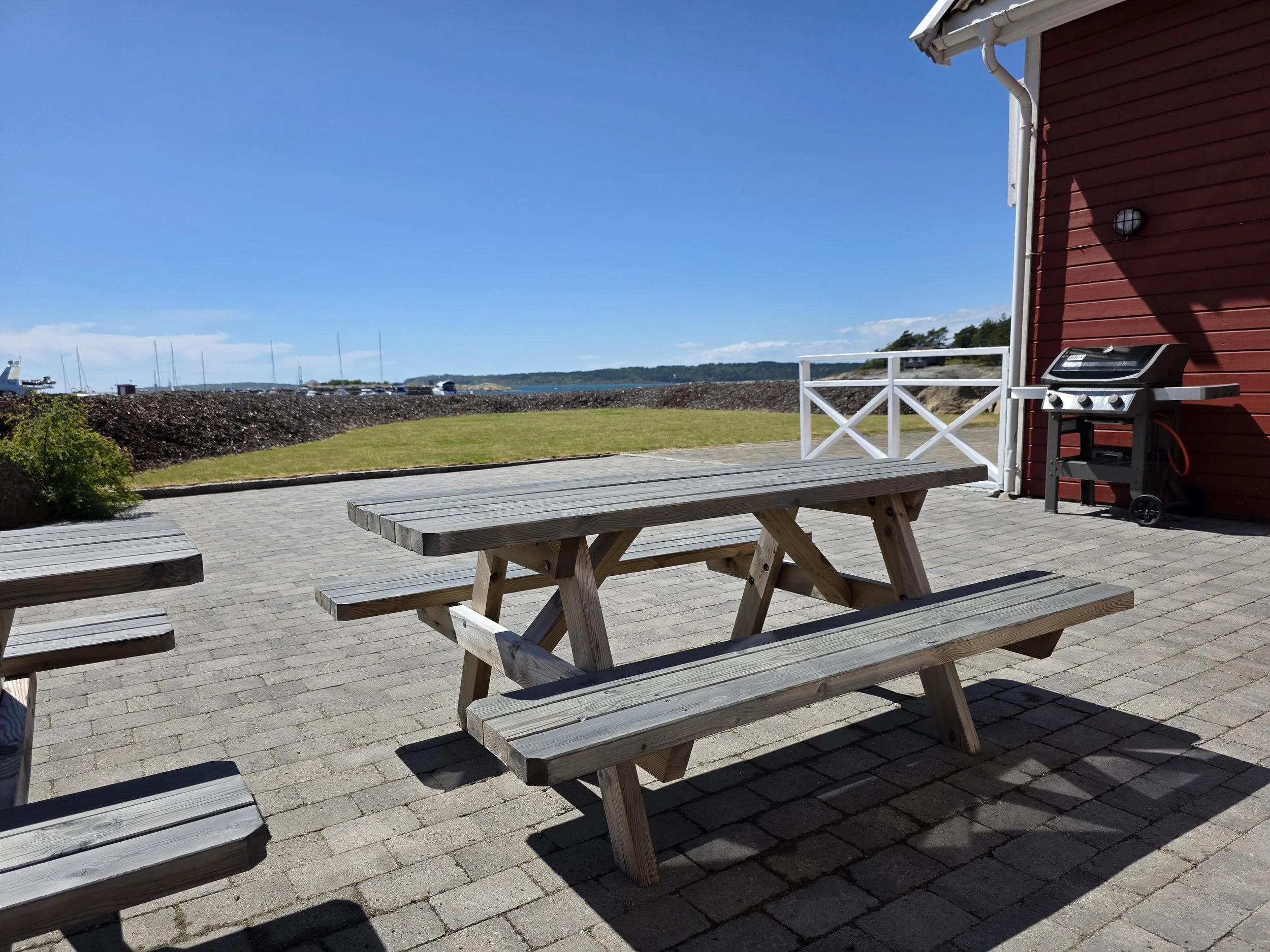 Utendørs terrasse med piknikbord, en grill, og en utsikt mot sjøen og båter i det klare været