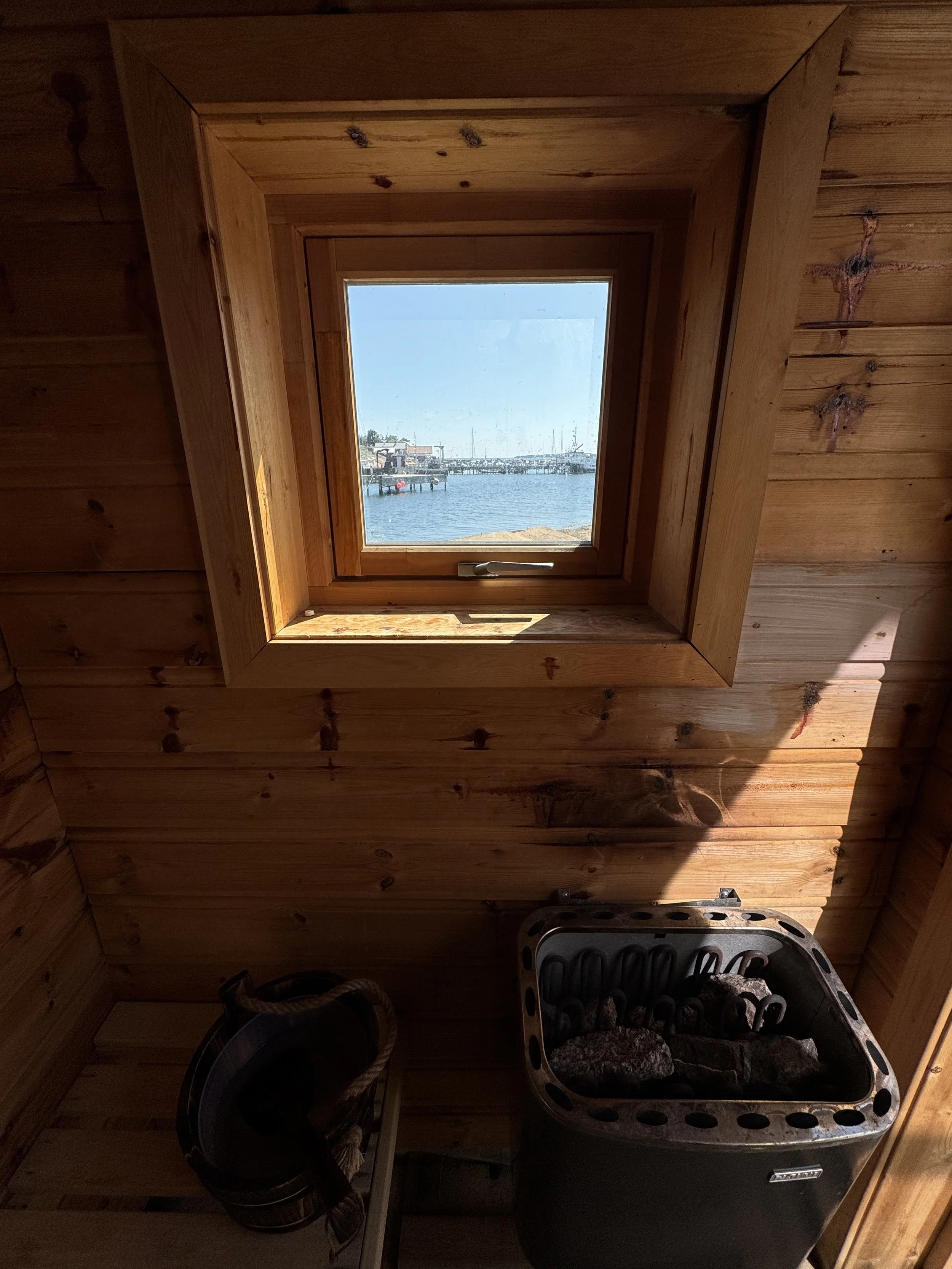 Innvendig i en trehus med et lite vindu som viser en sjøhavn med båter. Det er en badstue med en sauna ovn fylt med steiner, og en bøtte med håndkle i nærheten.