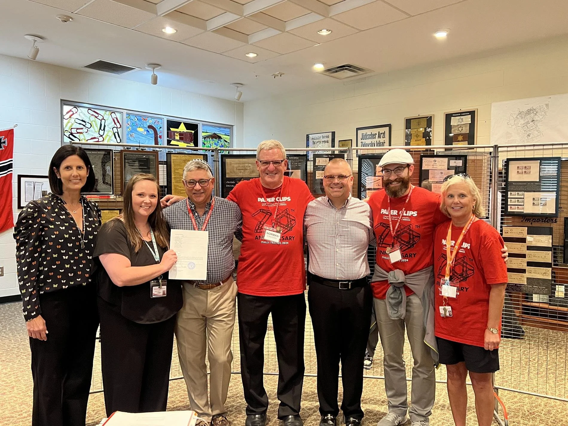 A group of six adults standing together in an indoor exhibit, some wearing matching red T-shirts and lanyards, with display cases and stained glass windows in the background.