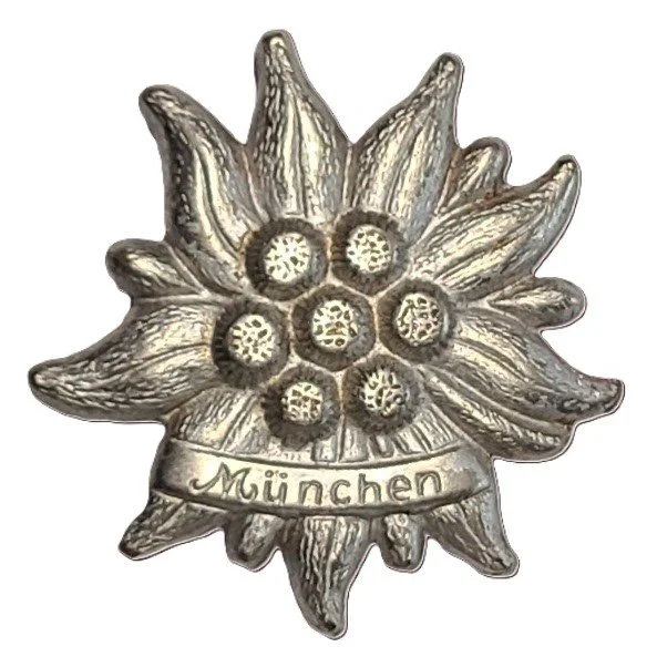 Silver-colored decorative medal or badge with a grapevine design and the word 'München' engraved on a banner beneath the grapes.