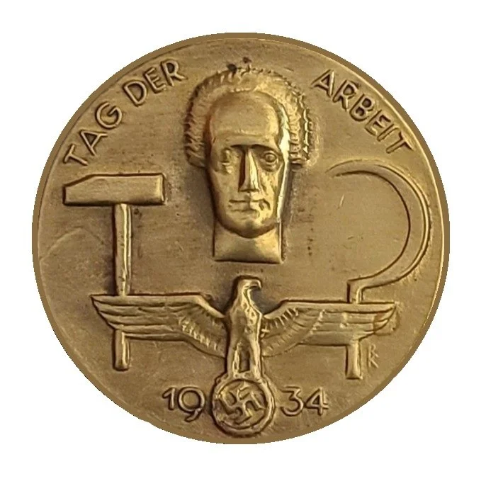 A gold-colored medal or coin with a face above an eagle holding a swastika inside a circle, and the words 'Tag der Arbeit' at the top. The date '1934' is at the bottom.
