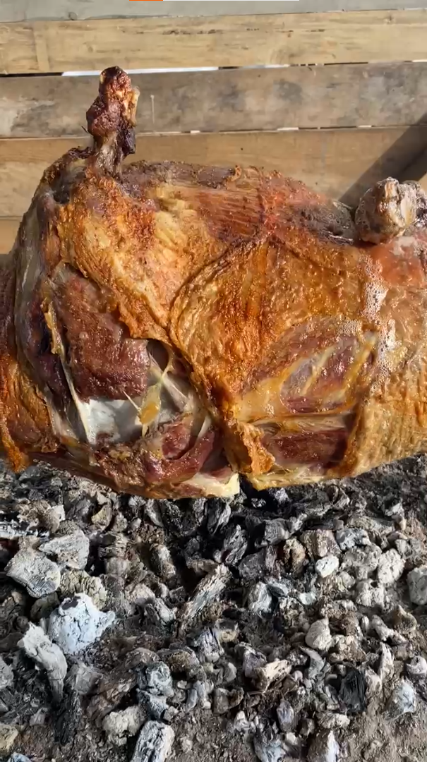 Cijelo pečeno janje odmara na vrućem uglju, sa zlatnom, hrskavom kožom, ispred drvene ograde.